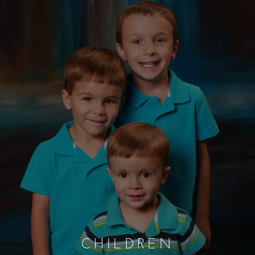 Three smiling children in teal shirts posed against a dark studio backdrop
