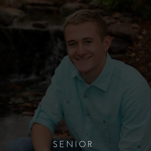 Smiling man in a light blue shirt sitting outdoors with a waterfall background, labeled “SENIOR”