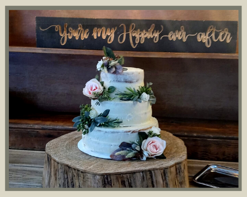 Three-tier white wedding cake with pink flowers on a wooden stump, under a sign reading “You’re my person and after”