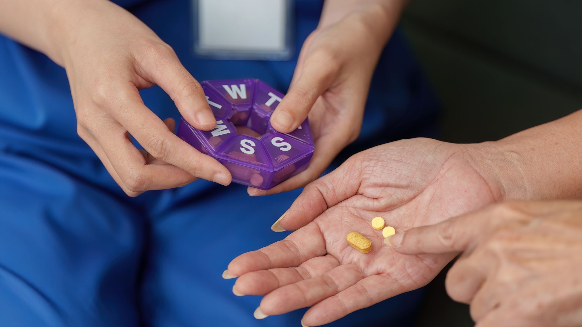 Hands holding a purple pill organizer and a palm with yellow tablets on blue clothing background