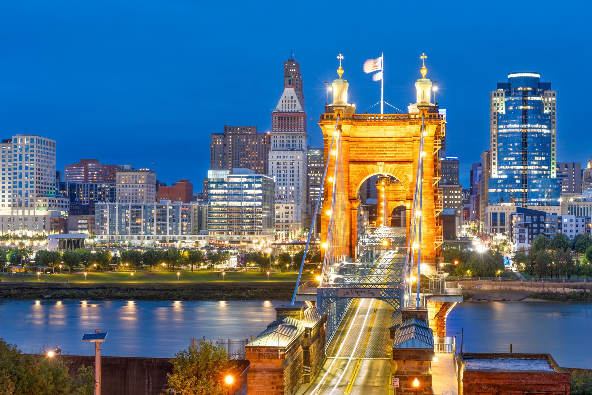 Lit-up suspension bridge at dusk over a river, with a city skyline glowing in the background.