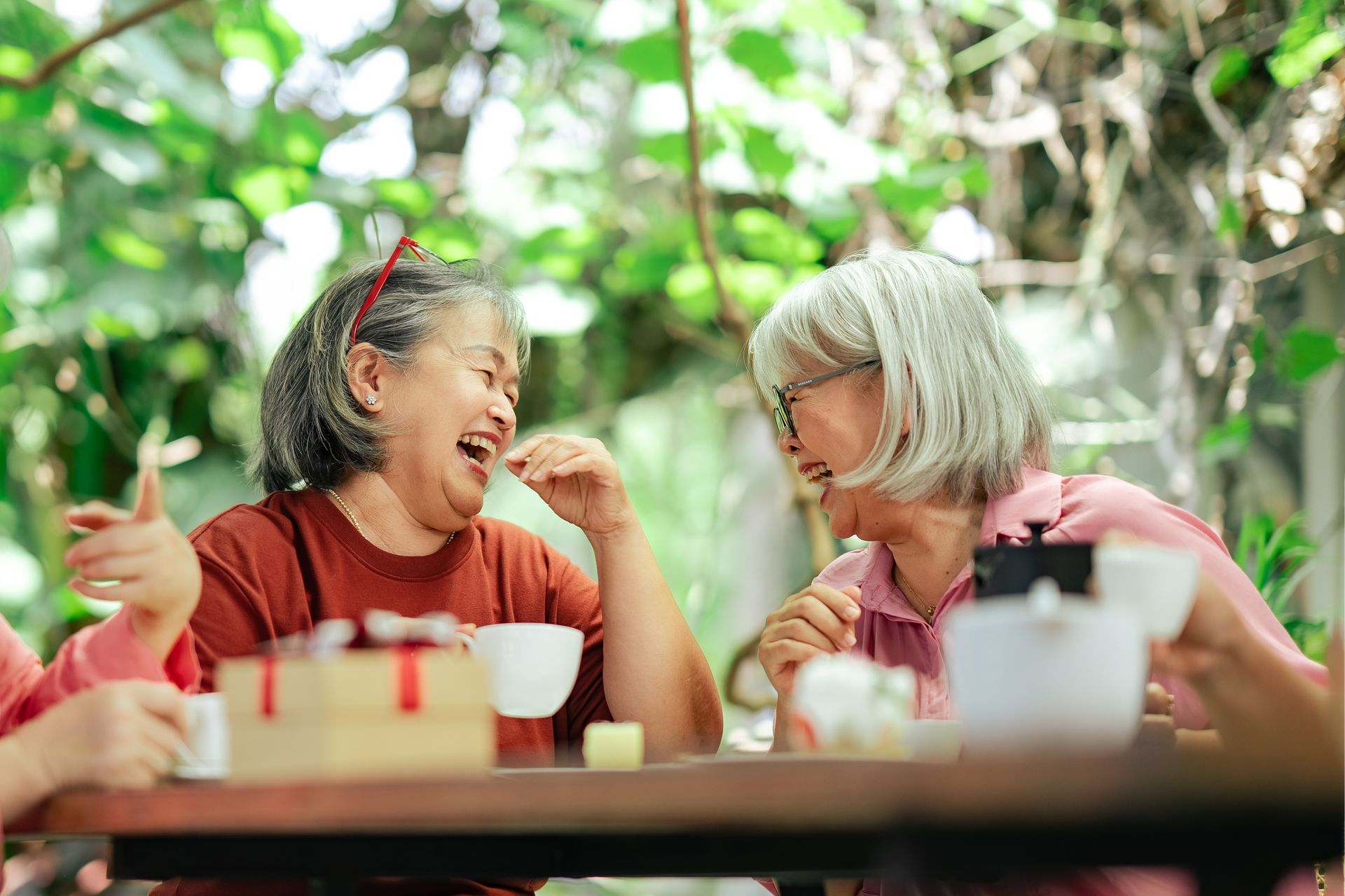 Two women laughing and chatting at an outdoor café table with drinks and greenery in the background