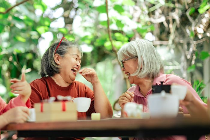 Two women laughing and chatting at an outdoor café table with drinks and greenery in the background