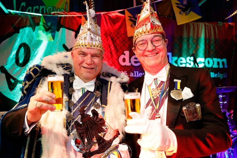 Twee mannen in kostuums toosten met bier voor een bord waarop staat kes.com