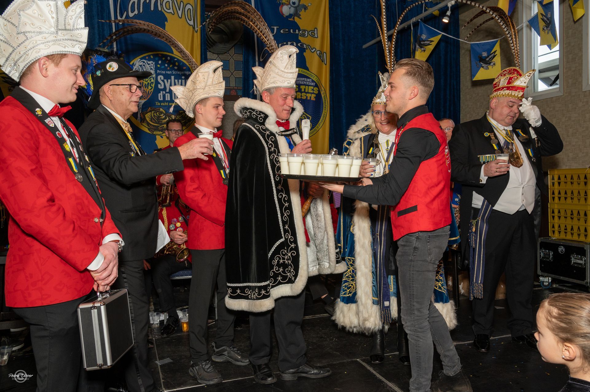 Een groep mannen in kostuums staat rond een man in een rood vest die een dienblad vasthoudt.