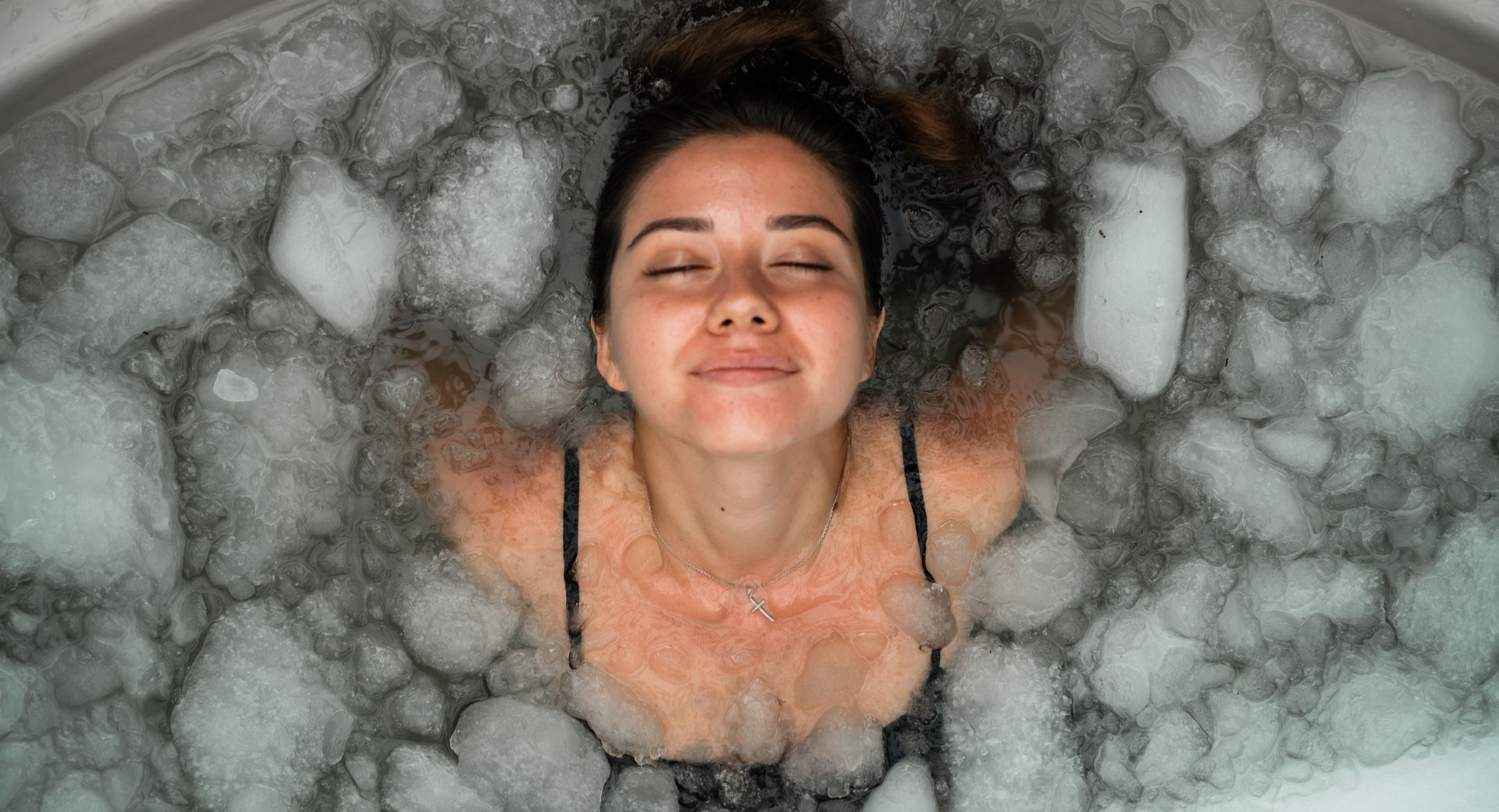 Woman submerged in ice bath, eyes closed, smiling, arms at sides. White ice surrounds her.