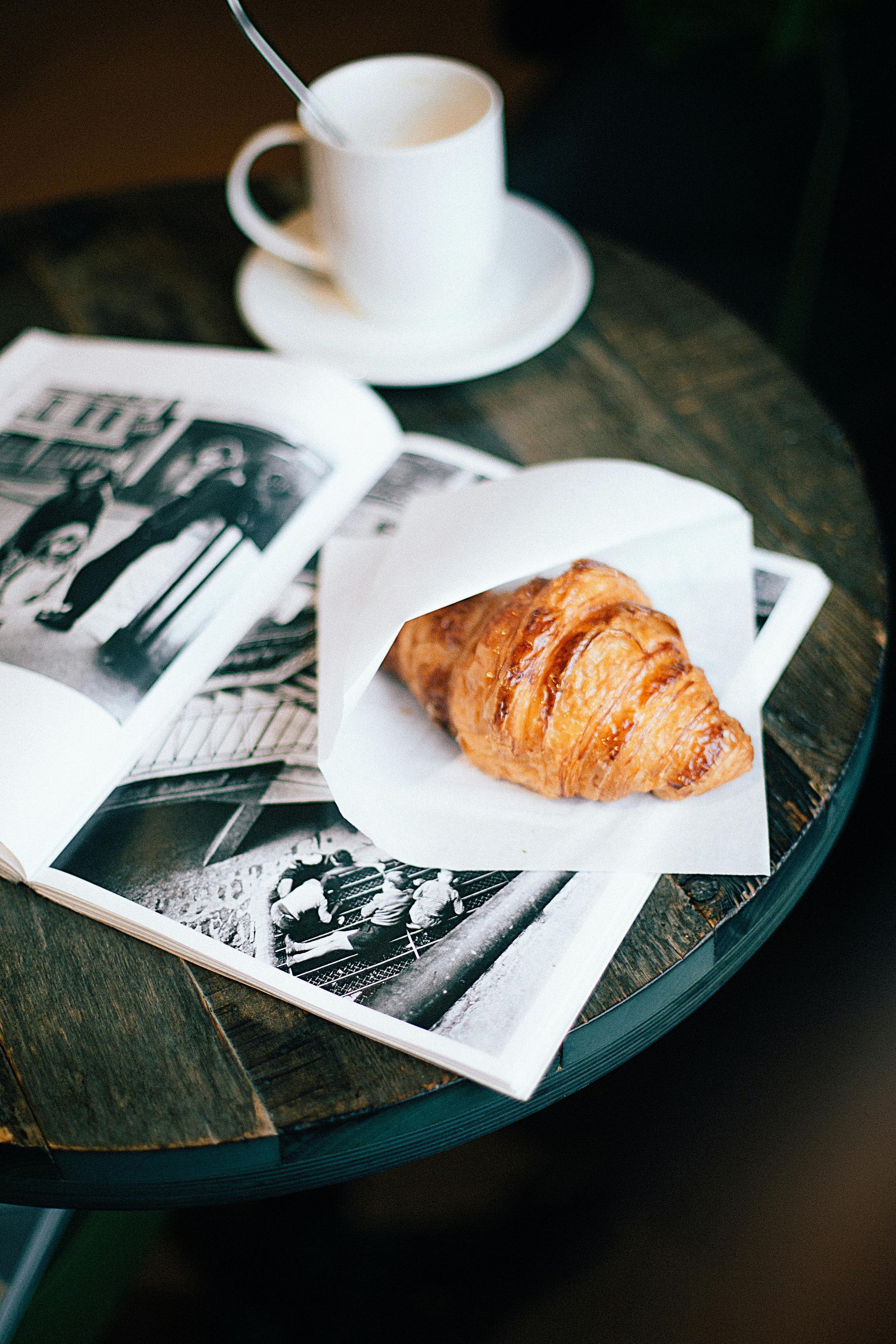 Croissant in paper, open book, and coffee cup on a dark wooden table.