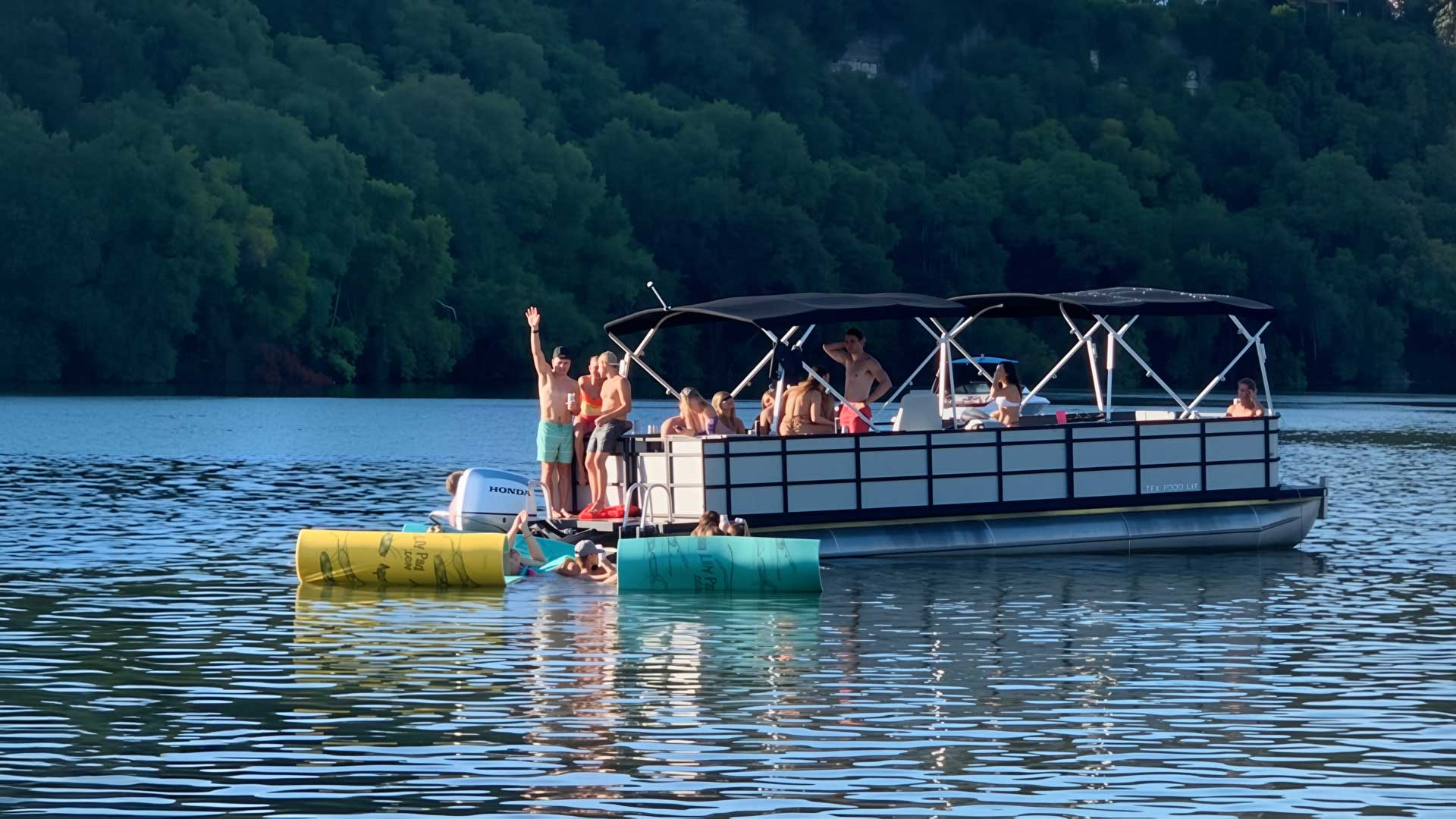 A pontoon boat on water with people on board; green trees and blue water in background.