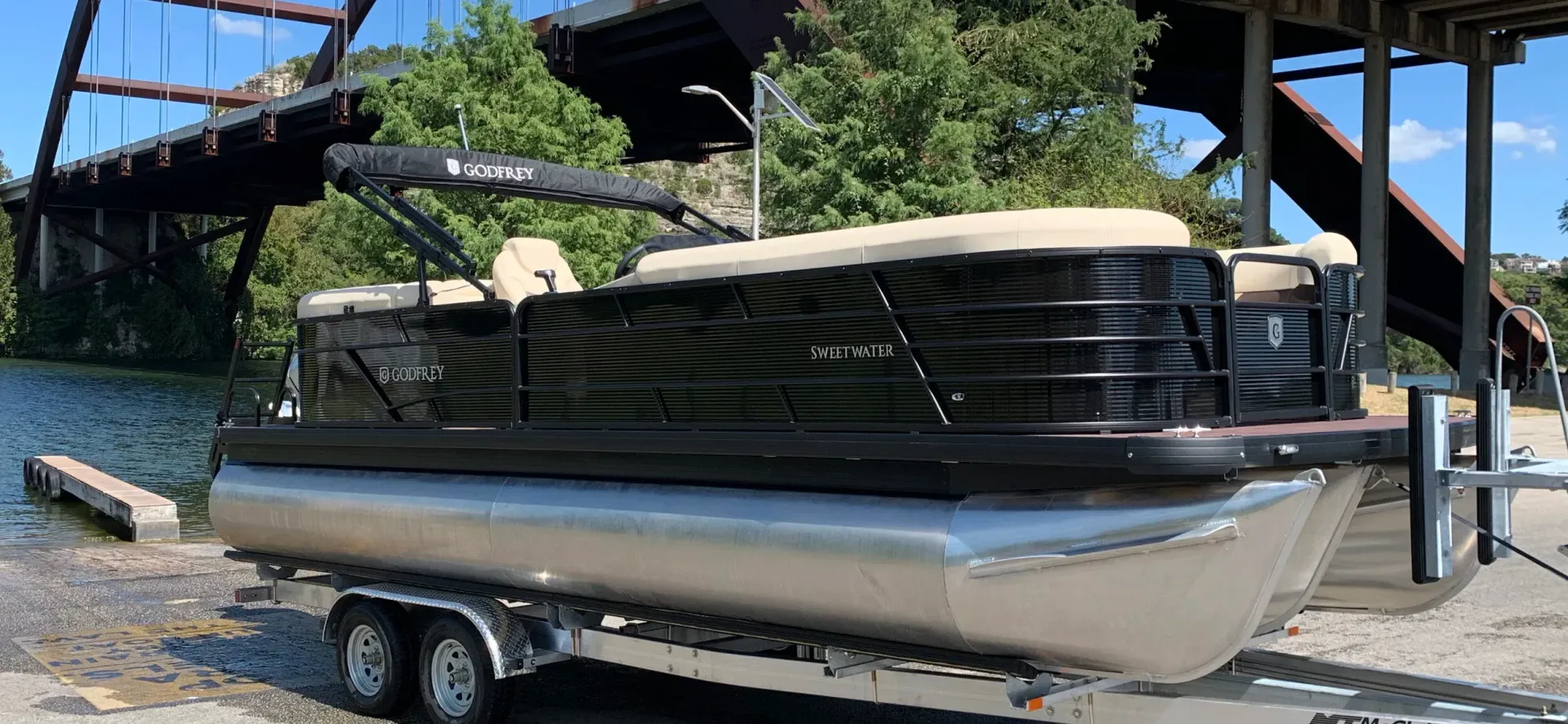 A pontoon boat on a trailer, under a bridge, with trees in the background.