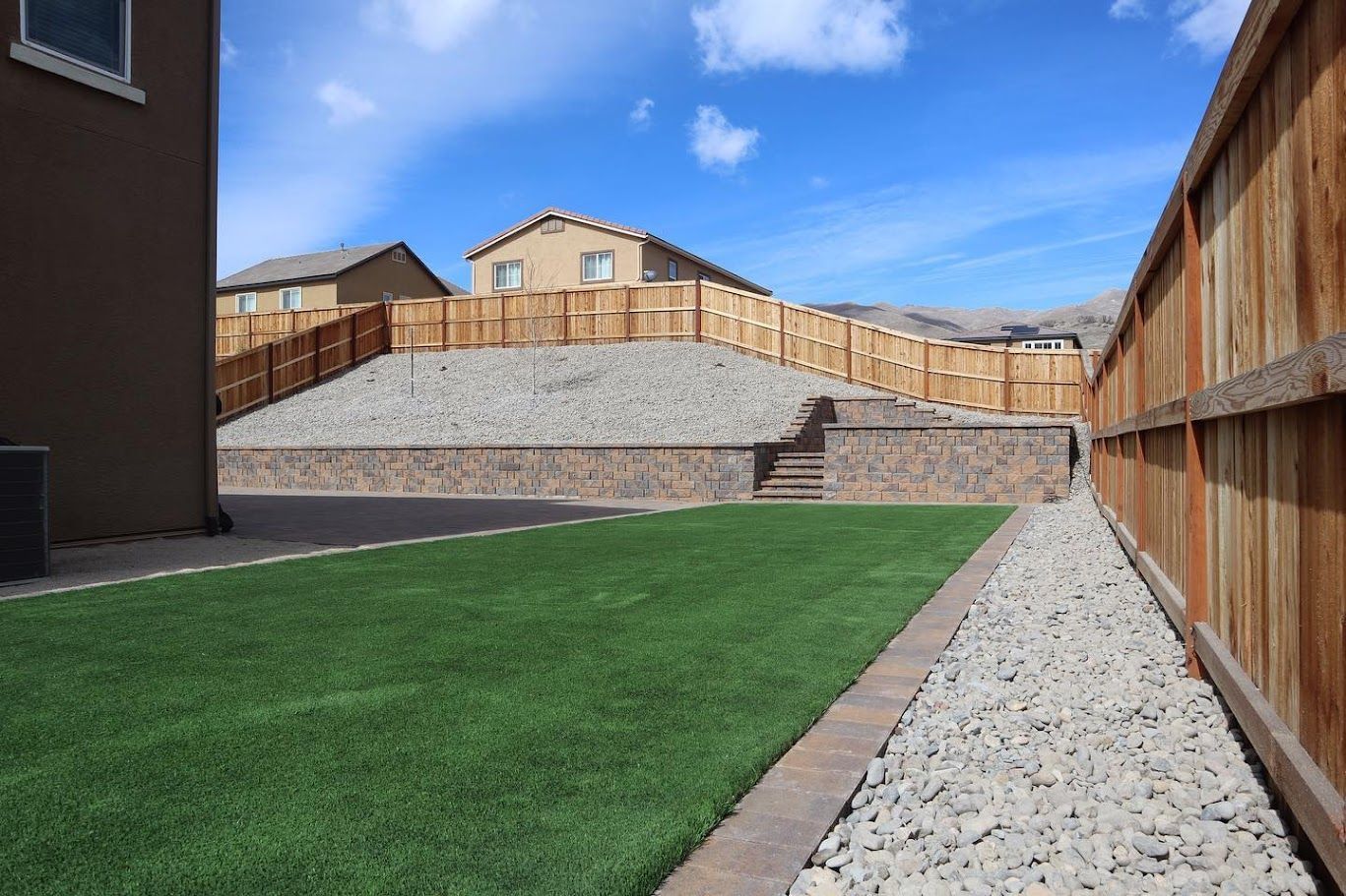 Backyard with green lawn, gravel paths, retaining wall, and wooden fences under a blue sky