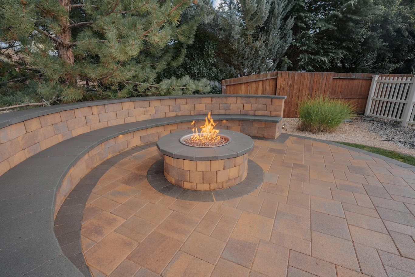 Curved stone fire pit with flames on a paved patio, surrounded by benches and a wooden fence