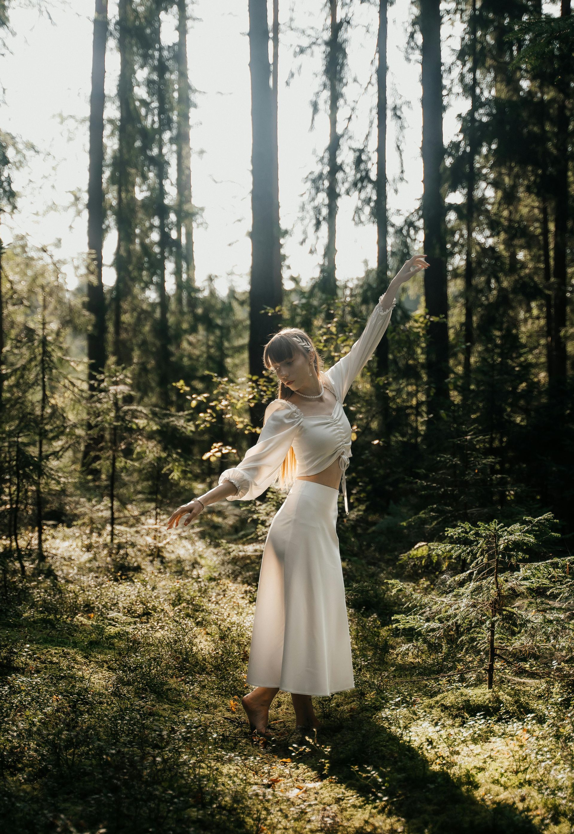 A woman in a white dress is standing in the middle of a forest with her arms outstretched.