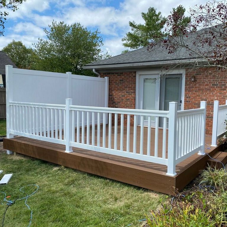 A white fence surrounds a wooden deck in front of a brick house.