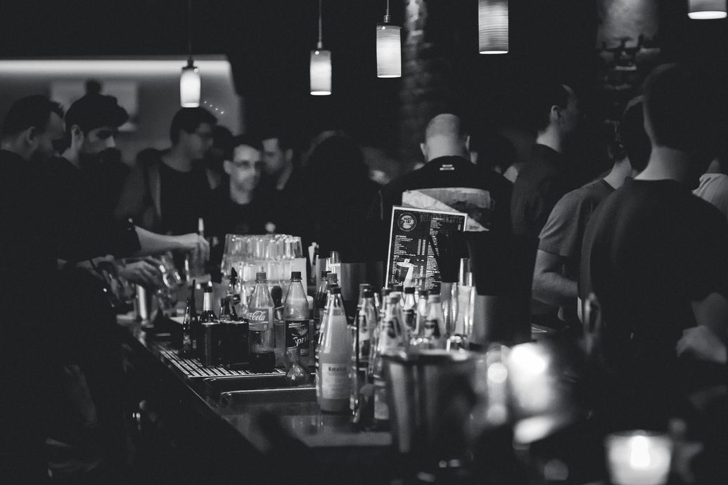Bar interior at night with people gathered, bartender behind bar with bottles and glasses.