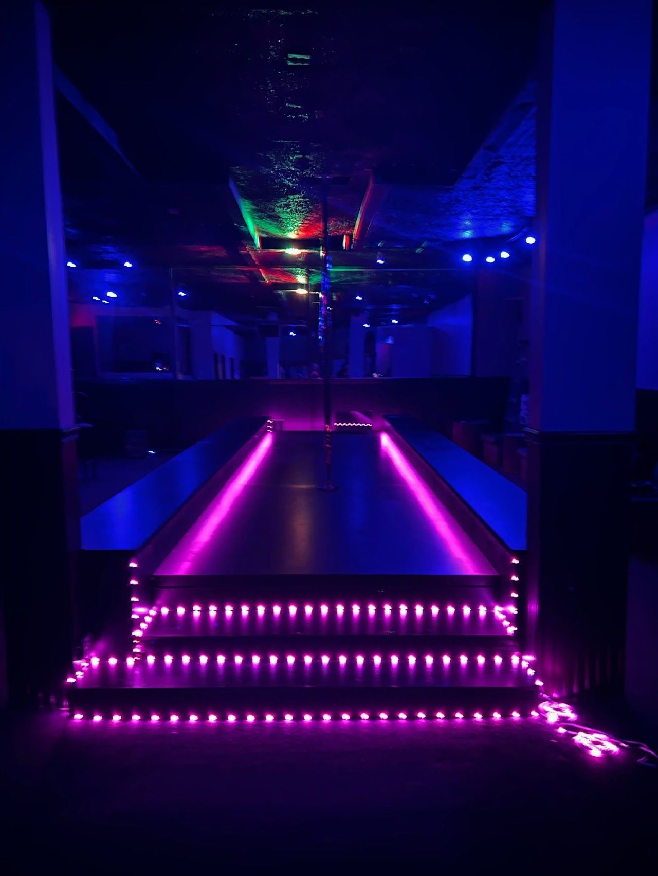 Purple and blue-lit nightclub entrance. Steps lead to a walkway with colored lights overhead, and empty seating areas.