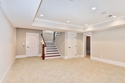 Empty beige basement room with white trim, recessed lights, and stairs leading upstairs