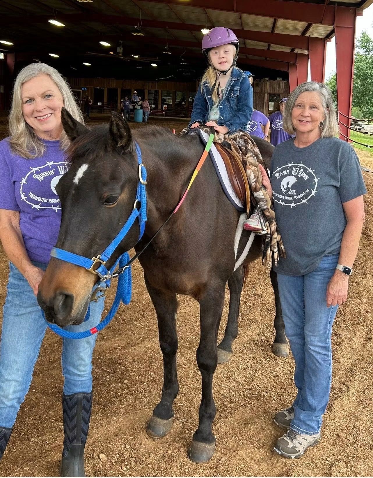 Child on a dark horse flanked by two women in a barn, all smiling and wearing helmets and jeans