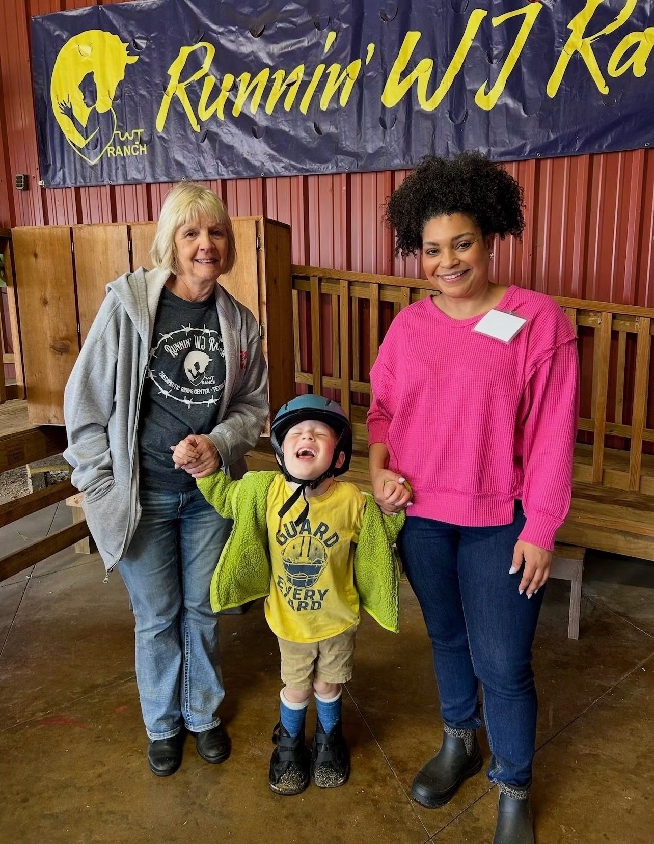 Three people standing indoors in front of a “Runnin’ WJ Ka” banner, smiling at the camera.
