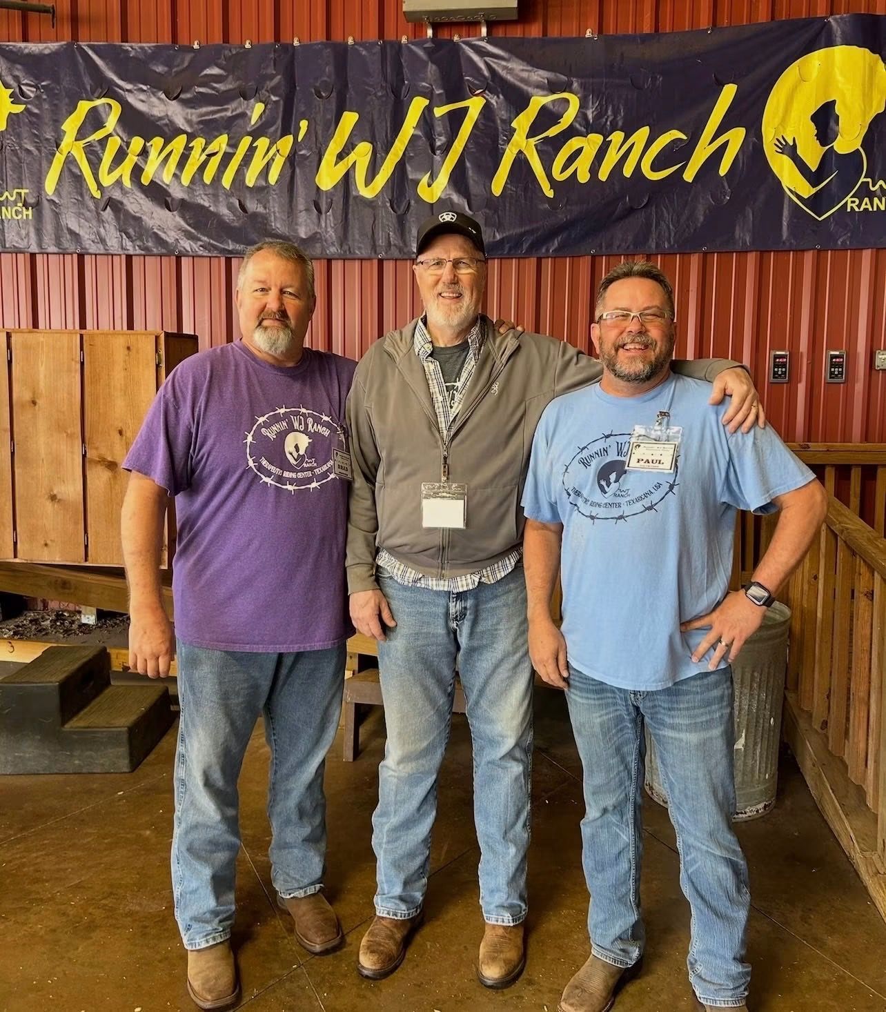 Three men pose smiling in front of a “Runnin’ WJ Ranch” banner and wooden fence.