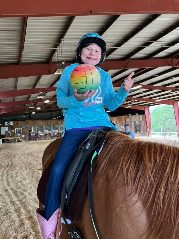 Person in a blue shirt and helmet rides a brown horse indoors, holding a rainbow-colored ball.