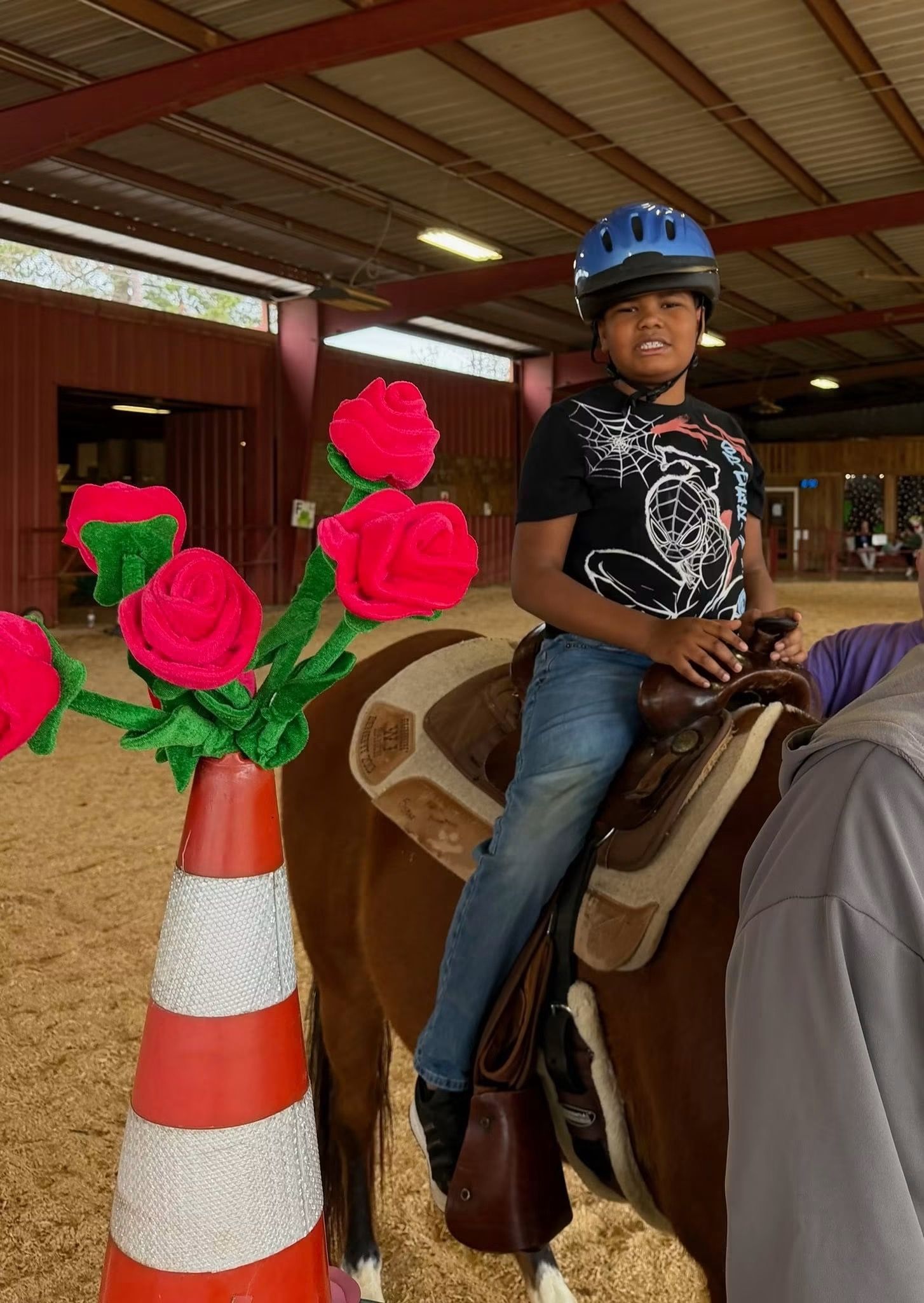 Child in a blue helmet sits on a brown horse beside a cone with bright pink flowers in an indoor stable