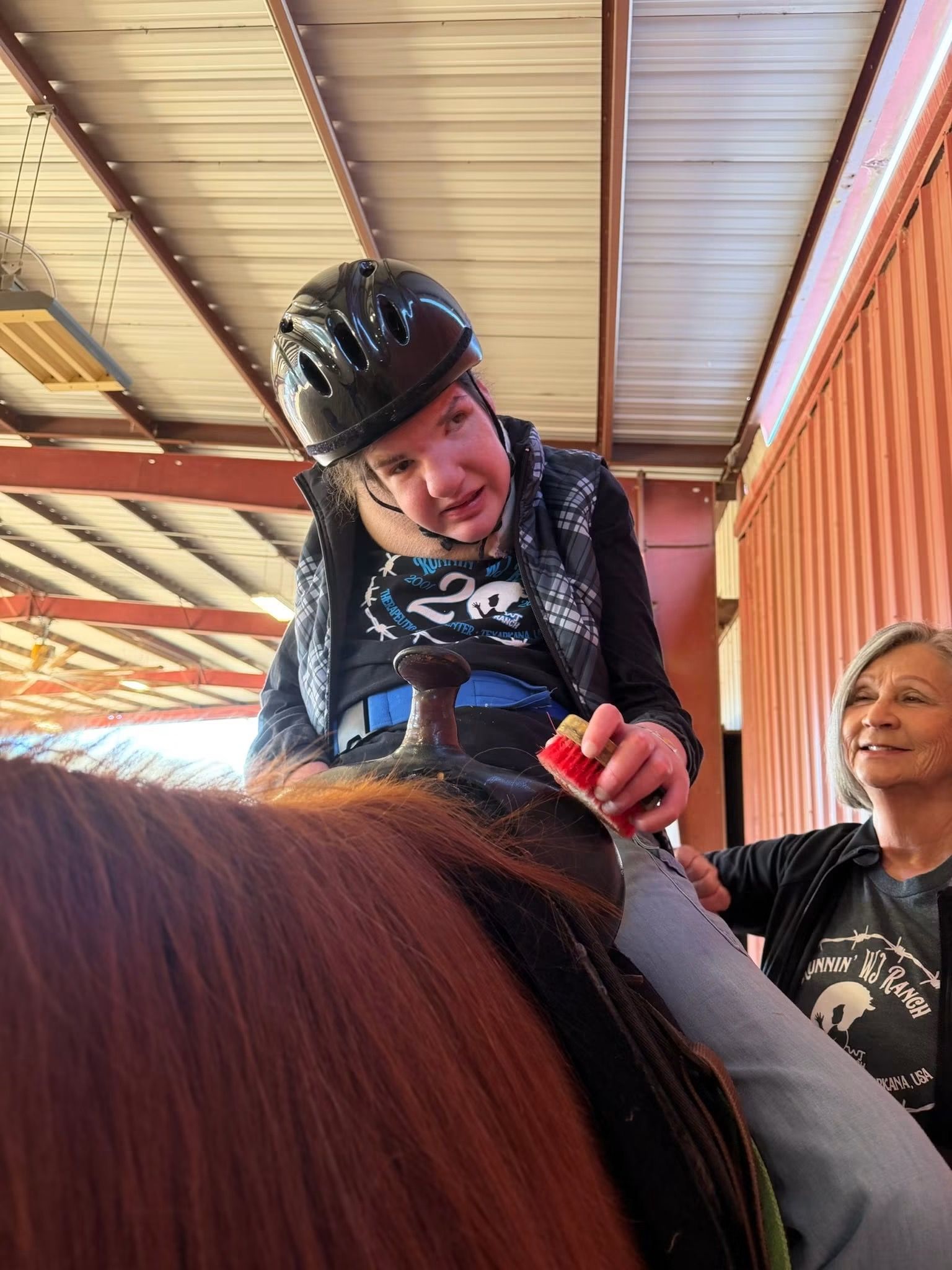 Helmeted rider smiling on a horse while another person steadies them under a metal-roofed shelter