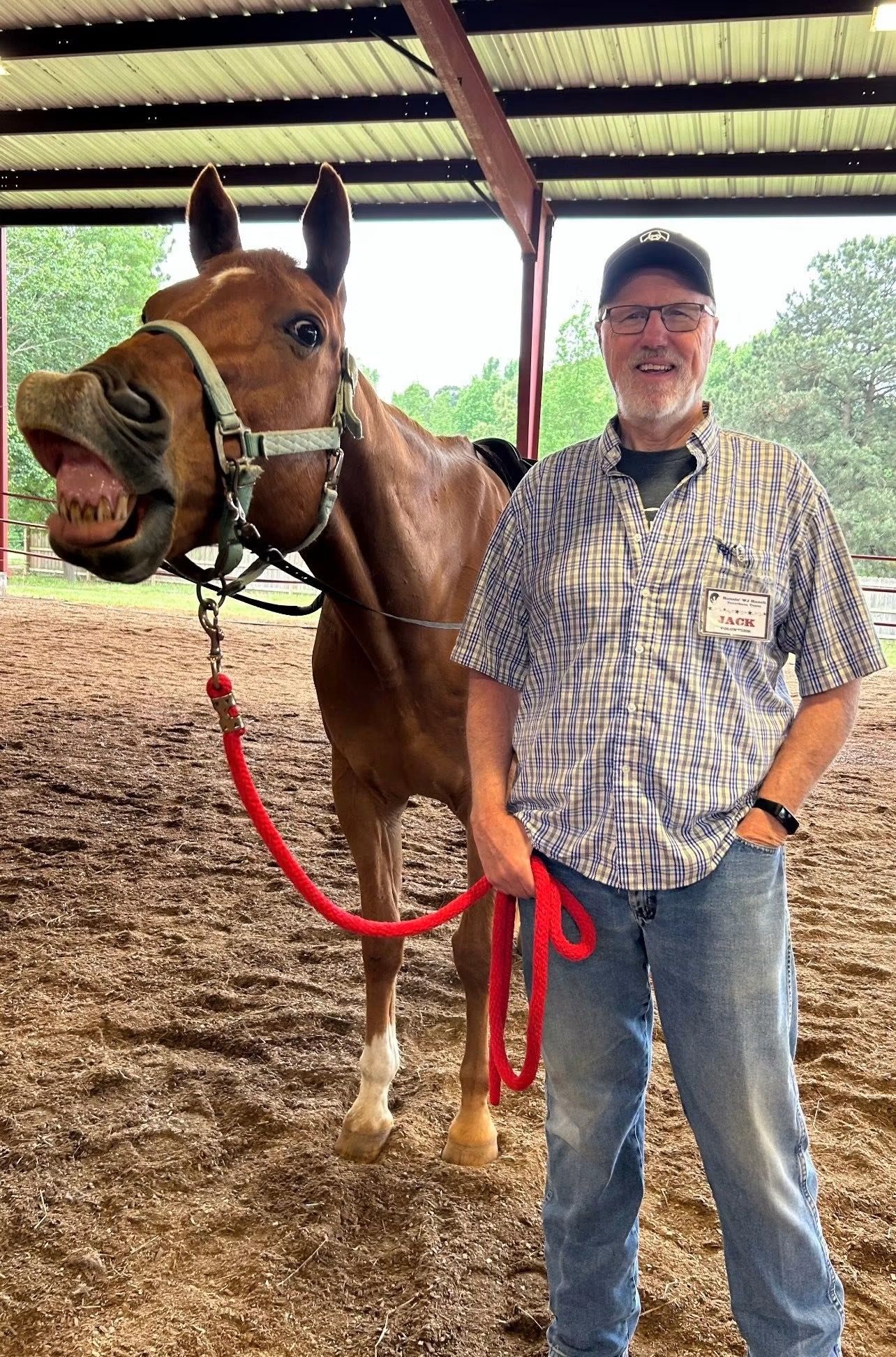 Man standing beside a saddled brown horse under a covered stable roof, both facing the camera.