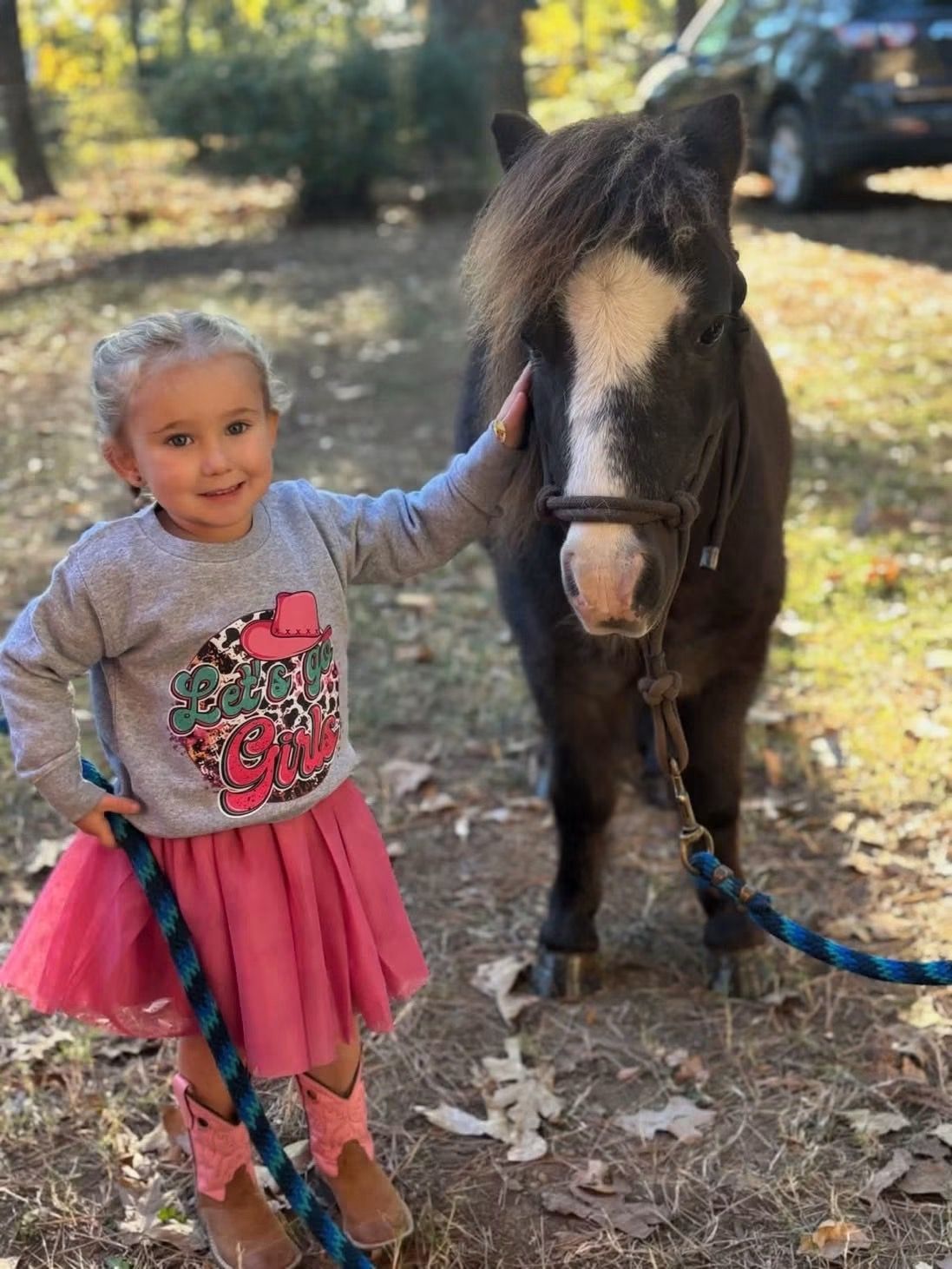 Young child smiling beside a small horse outdoors, with a pink skirt and a blue lead rope