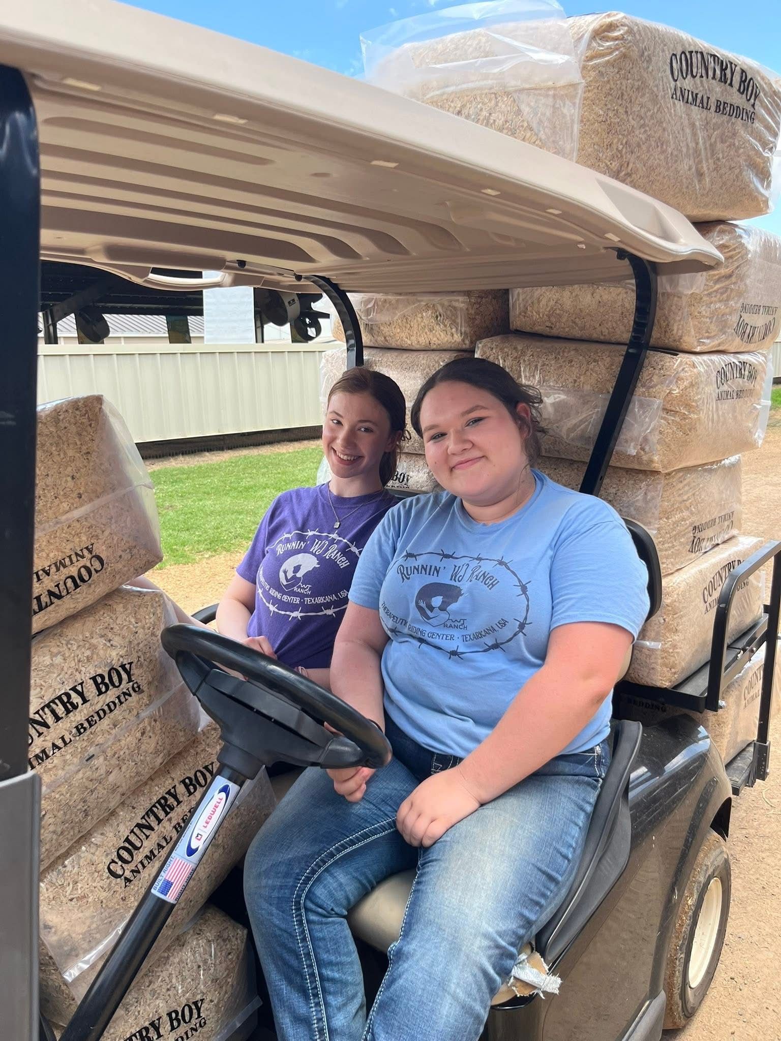 Two people sitting in a golf cart, surrounded by stacked cardboard boxes in a warehouse-like setting.