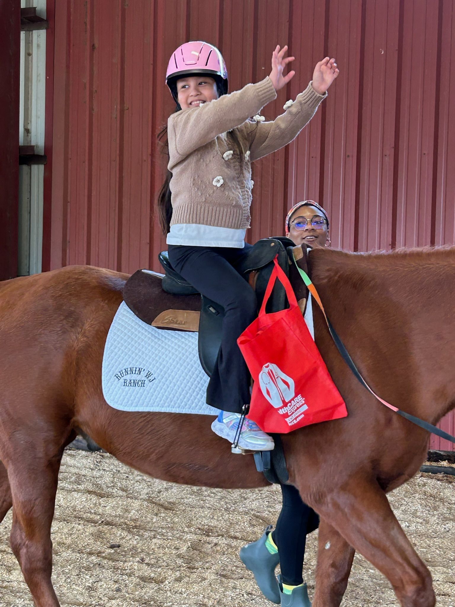 Child wearing a pink helmet rides a brown horse, arms raised, in a barn arena.