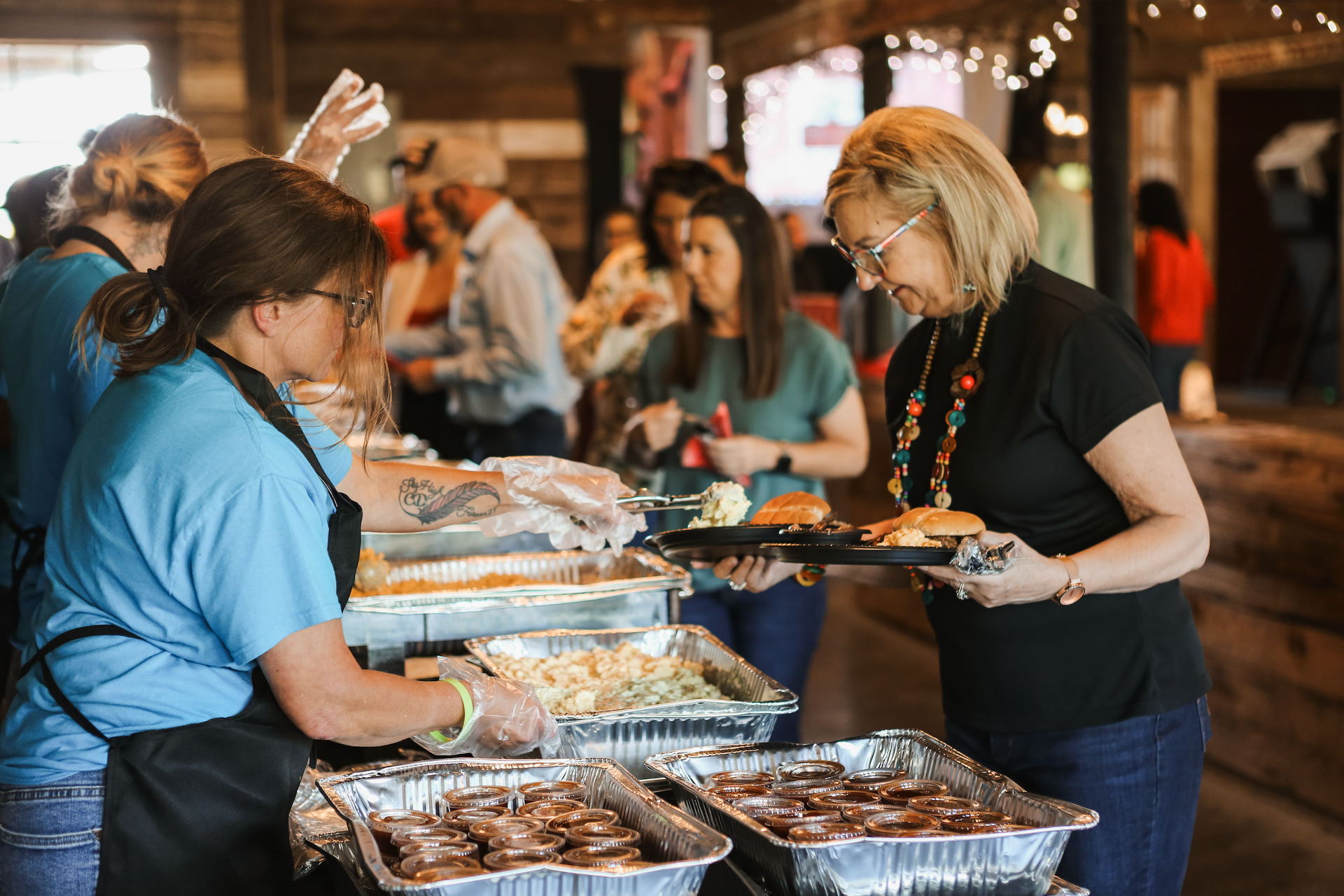 People serving buffet dishes at a crowded indoor event, with trays of food on a long table