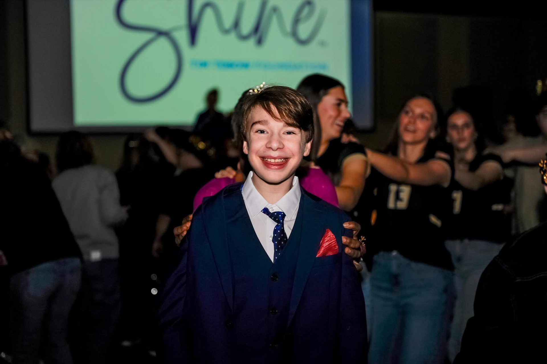 Smiling person in a navy suit stands in front of a screen reading “Shine” at an event