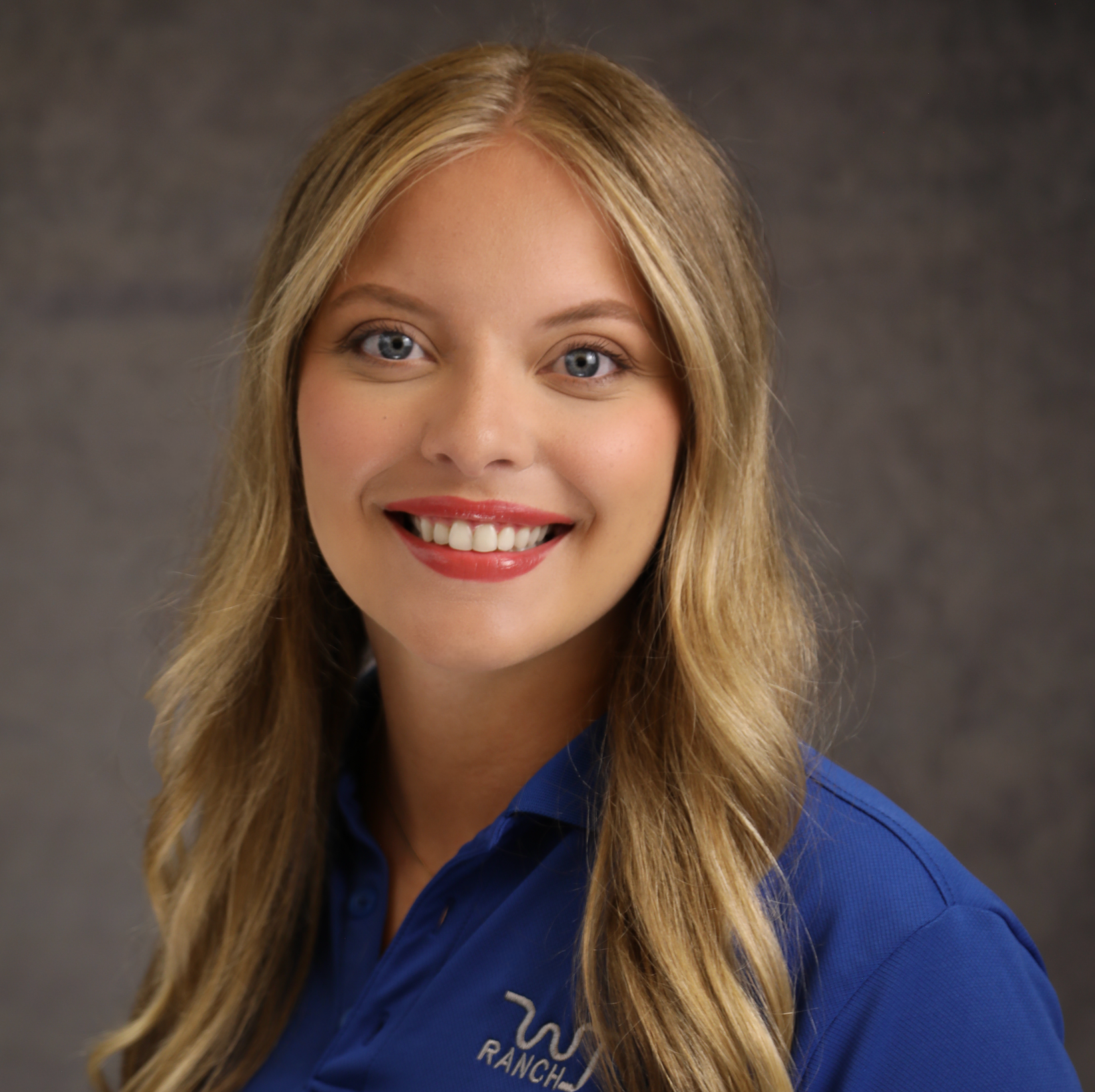 Smiling blonde woman in a blue polo shirt against a gray studio backdrop