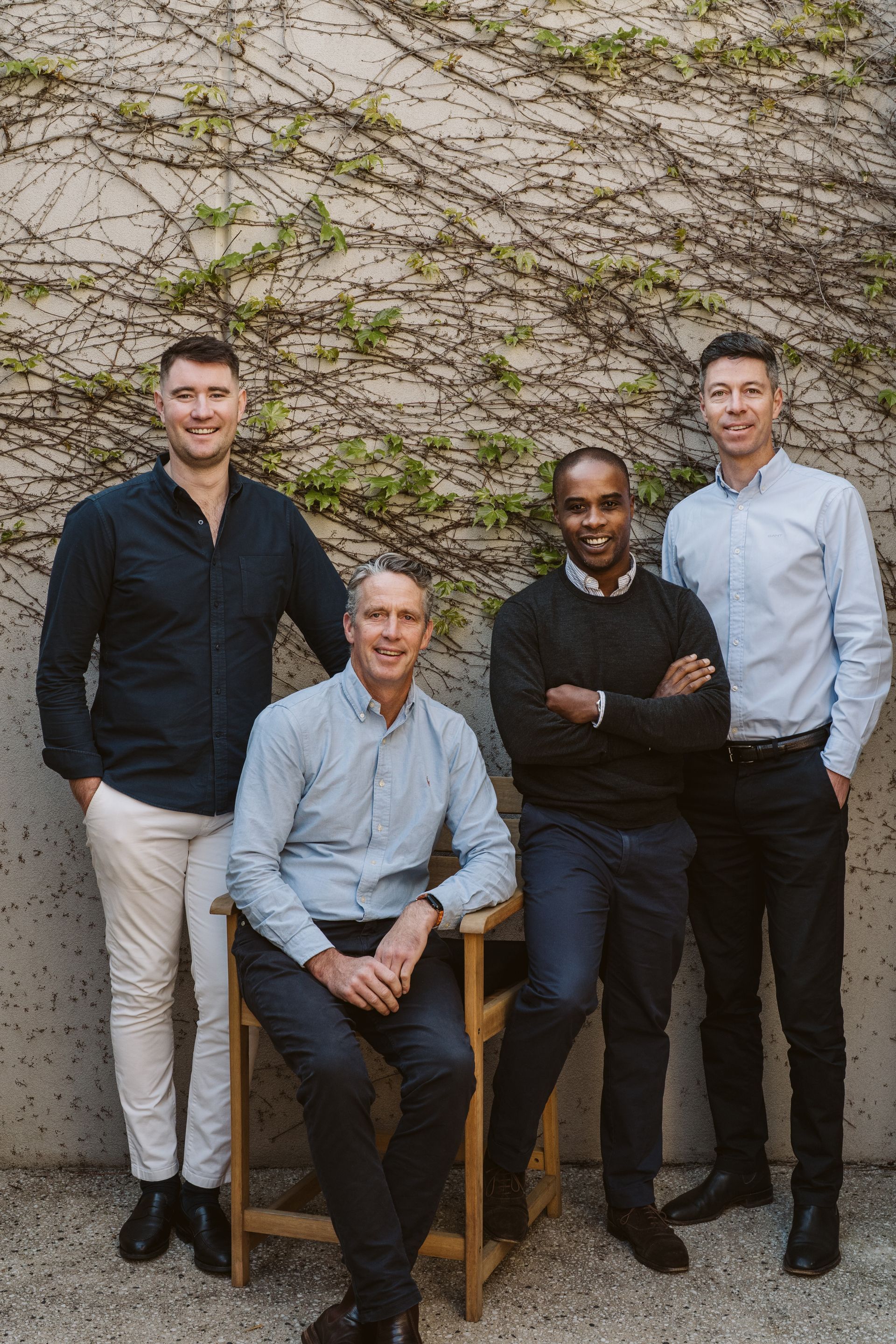 Four men pose together in front of a wall with ivy. Two stand, two sit, all smiling.