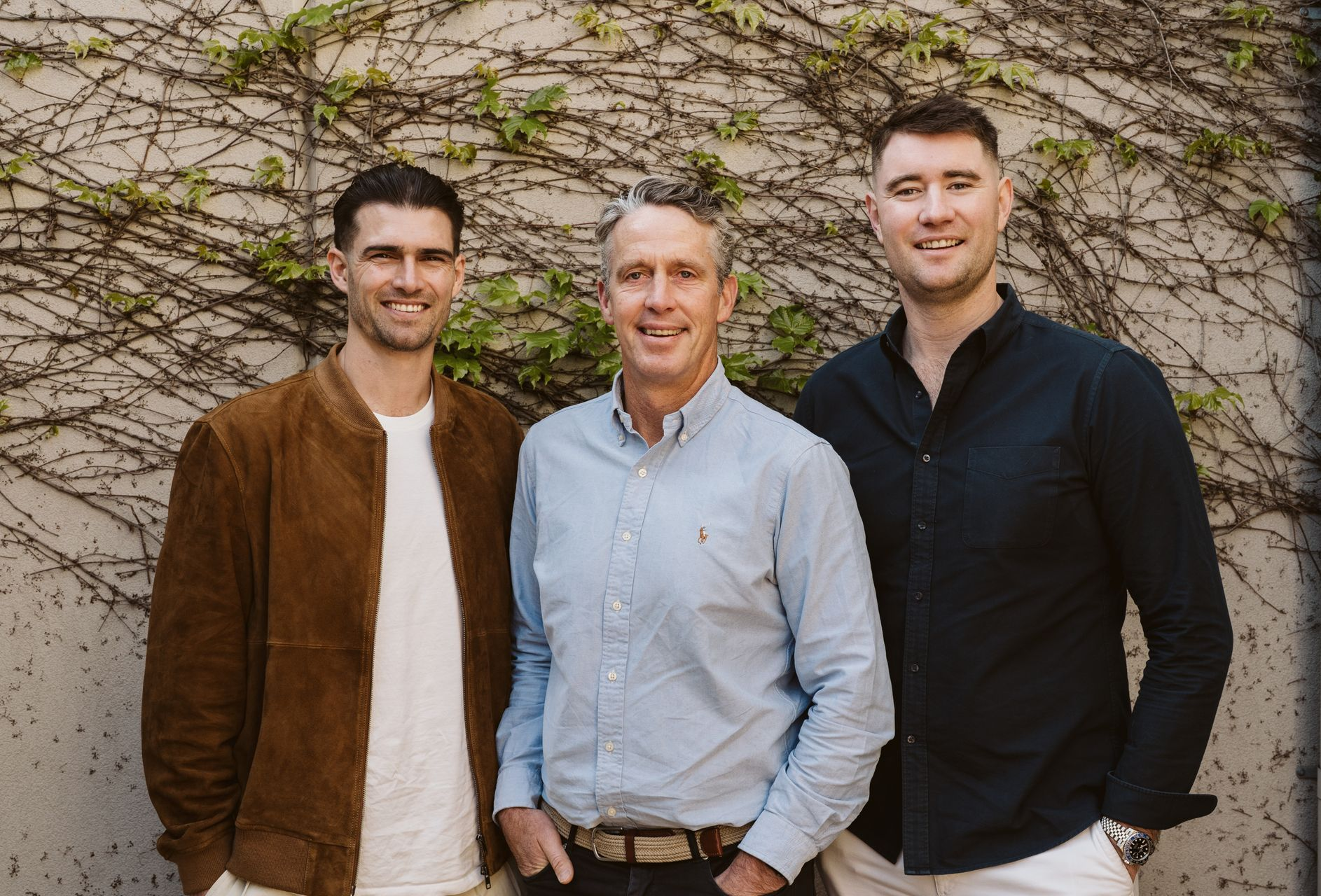Three men standing in front of a plant-covered wall. One in a brown suede jacket, one in a blue shirt, one in a dark shirt.