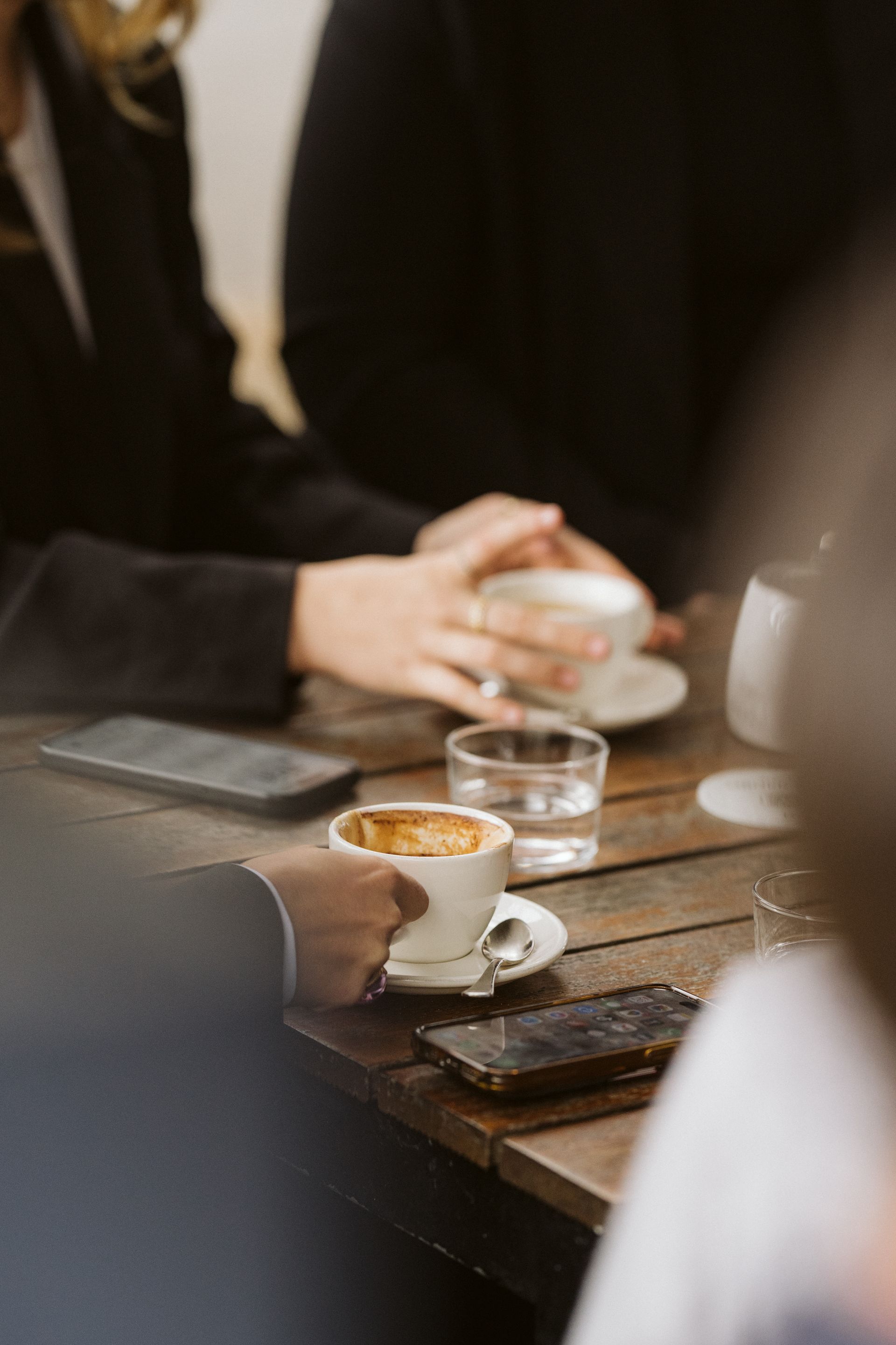 People at a table holding coffee cups, smartphones, and a glass of water. Wooden table, indoors.