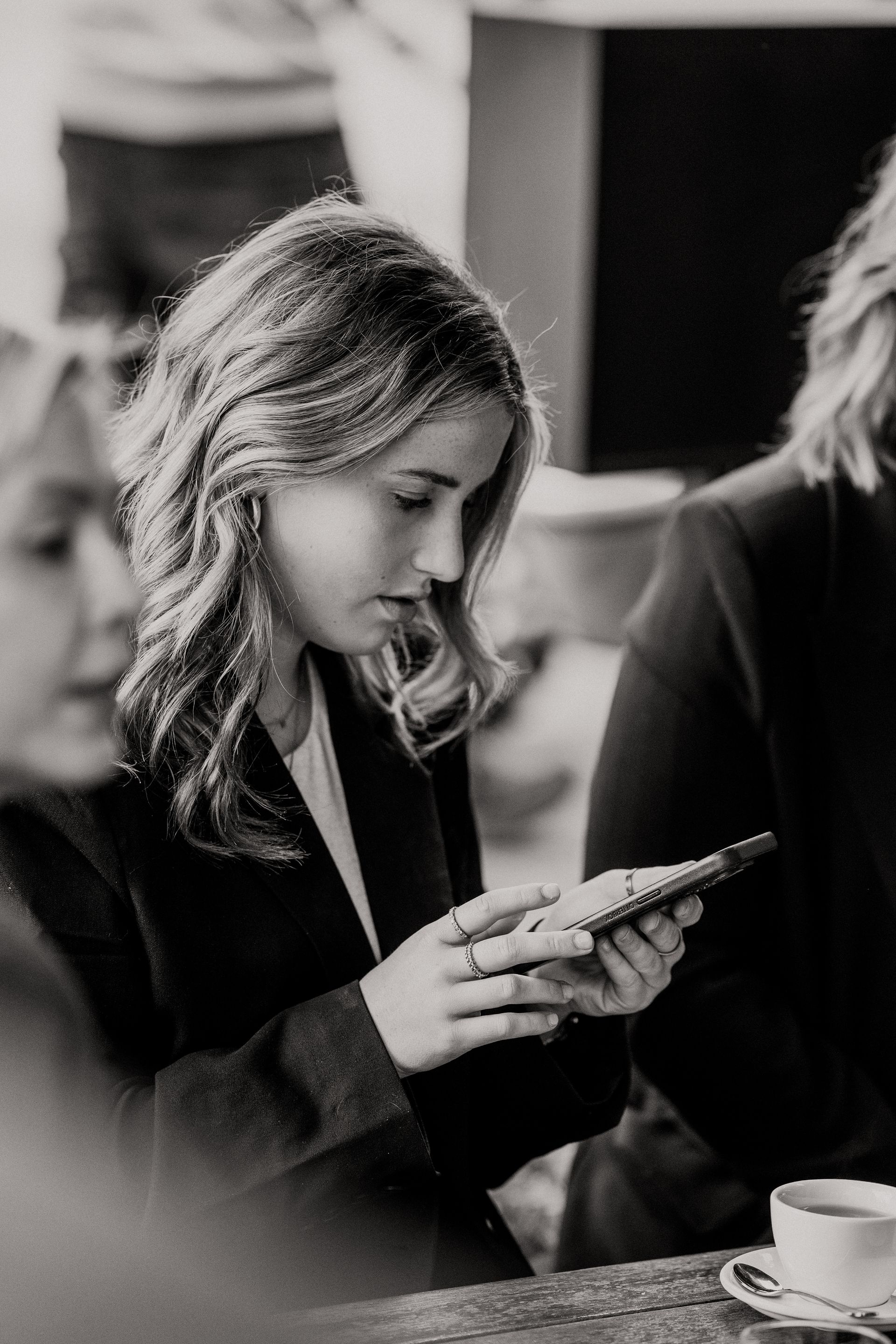 Woman in blazer looks at a phone; seated at a table with others, in black and white.