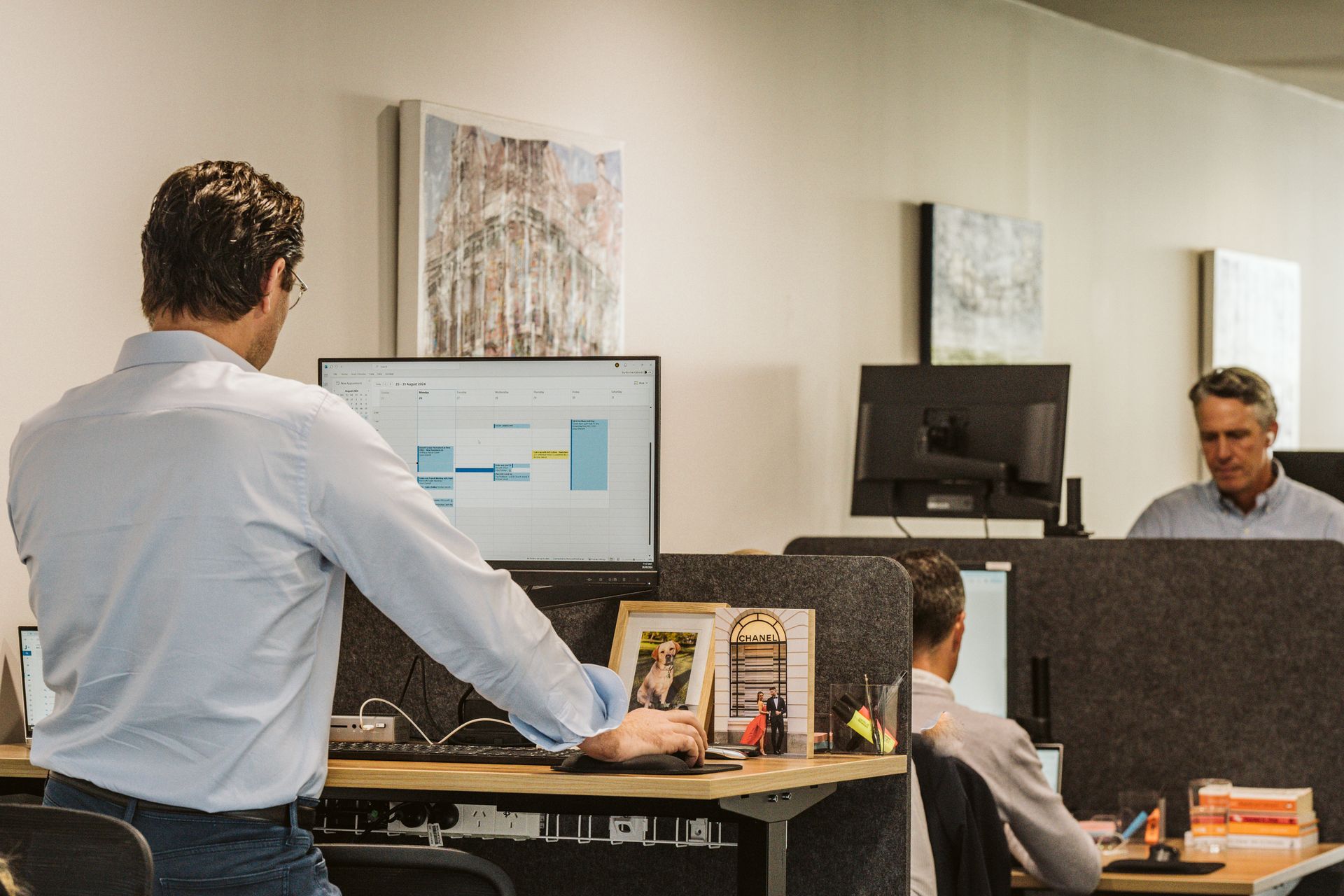Man standing at standing desk in office, looking at computer screen with two other men working in the background.