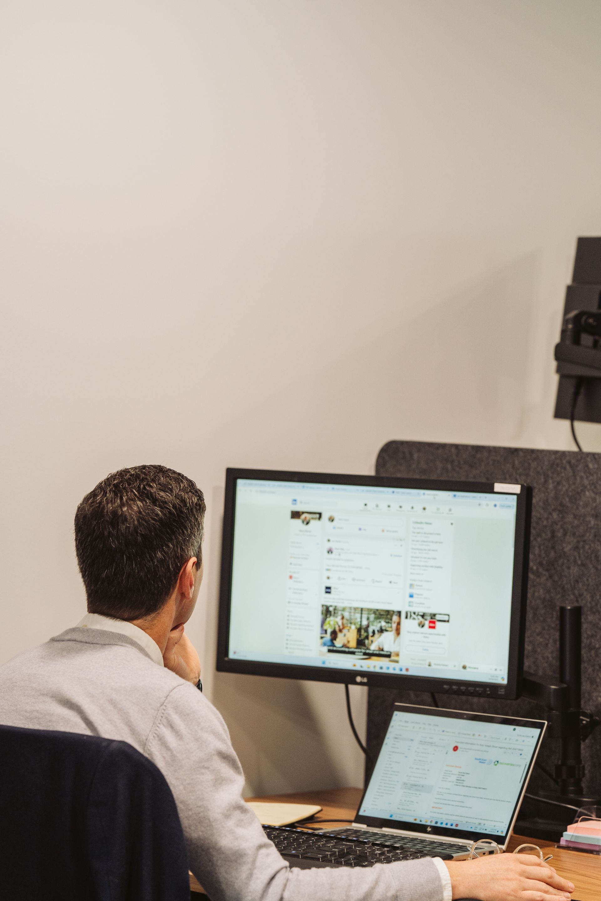 Person seated at a desk, facing a computer monitor and laptop. They are looking at the screens, hand on their chin.