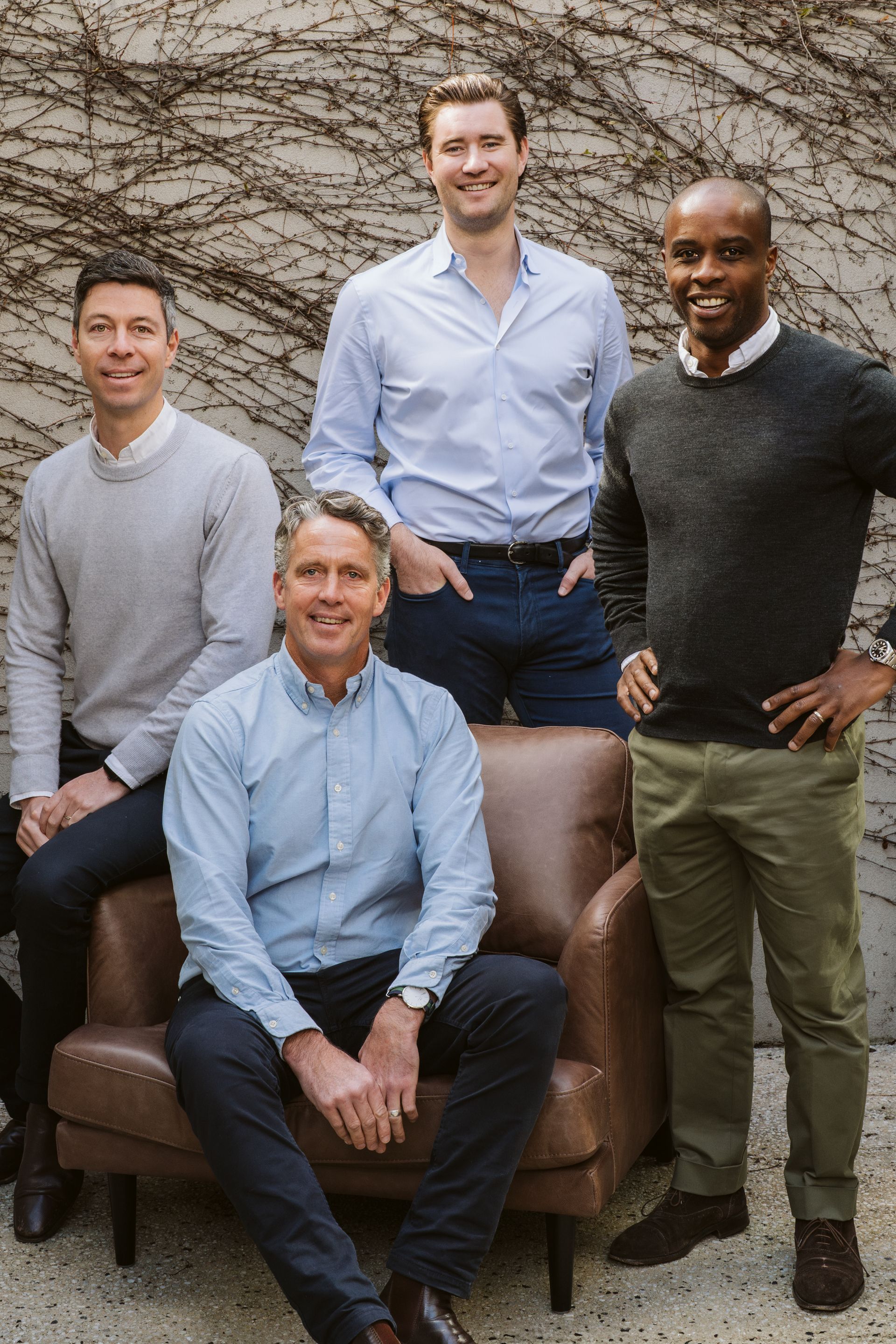 Group of four men smiling, posing near a brick wall, one seated in a leather chair.