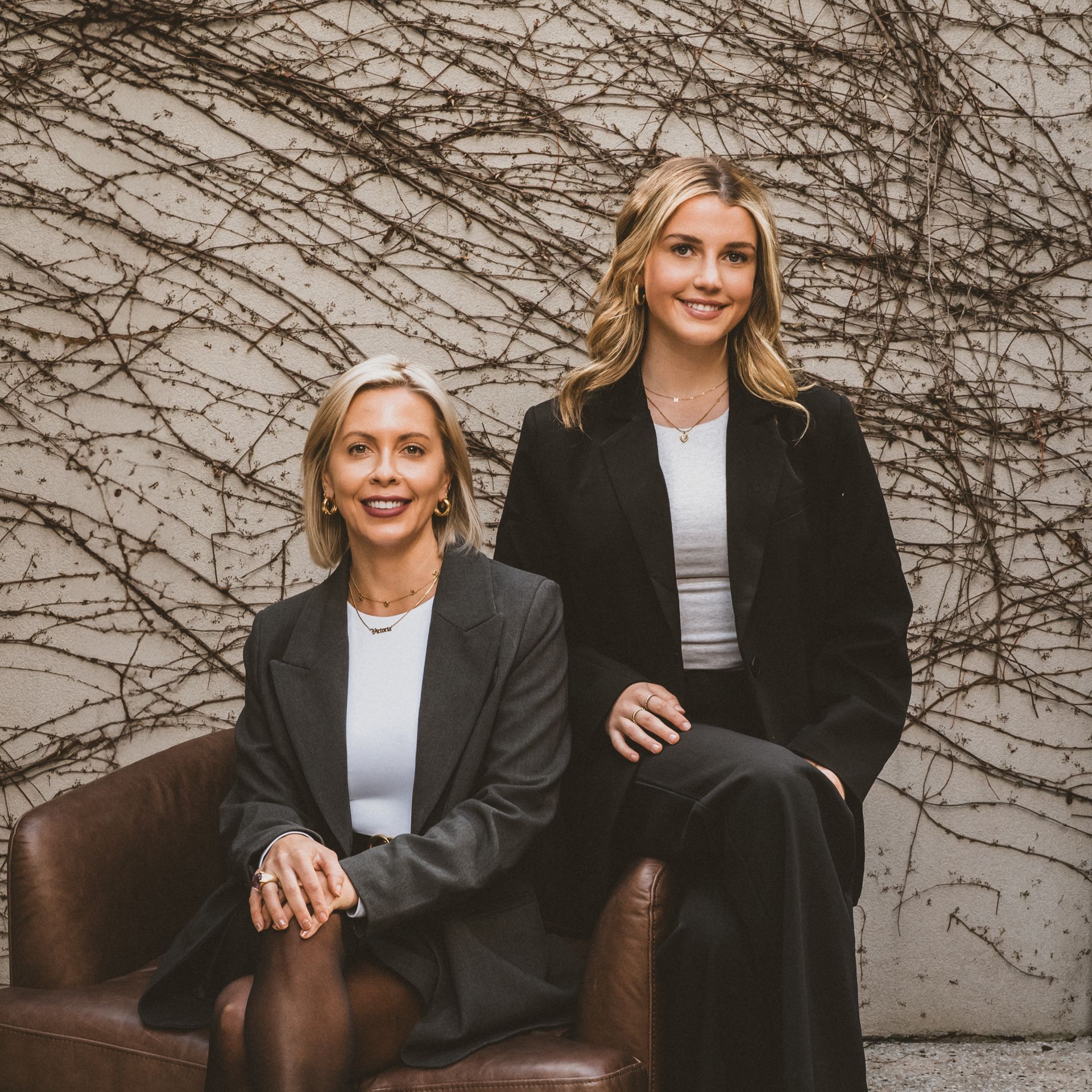 Two women in business attire pose in front of a wall covered in vines.