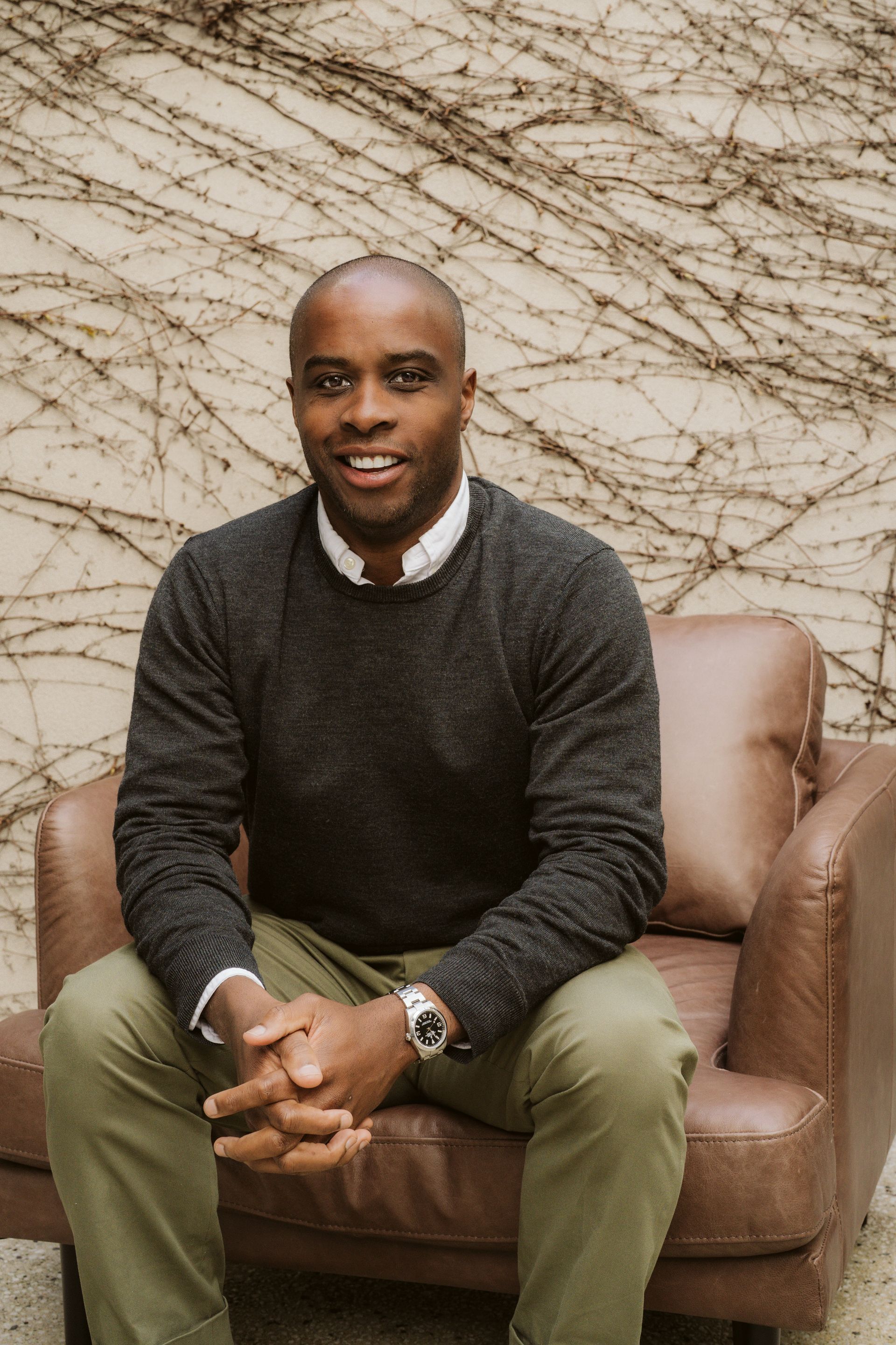 A smiling man sits in a brown chair, hands clasped, wearing a sweater and green pants. Vines grow in the background.