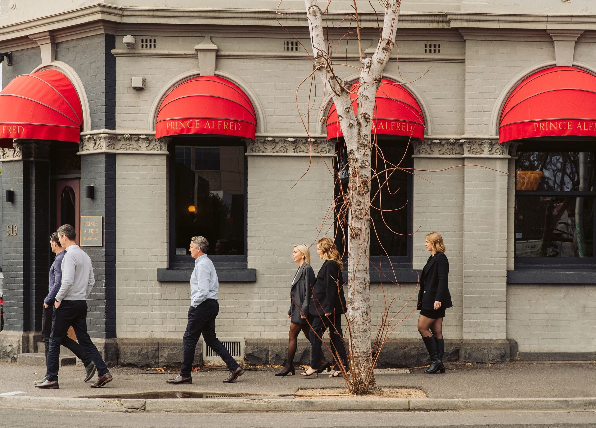 People walking on a sidewalk in front of a grey building with red awnings and large windows.