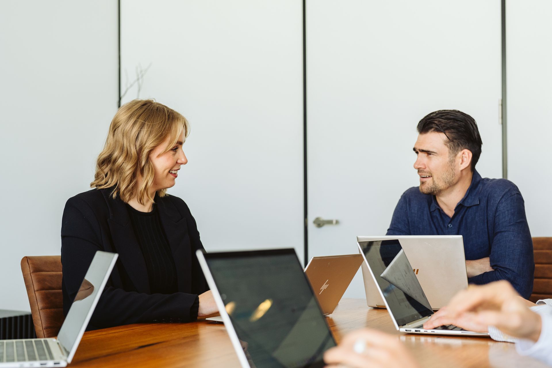 Two people conversing at a table with laptops in a bright office setting.