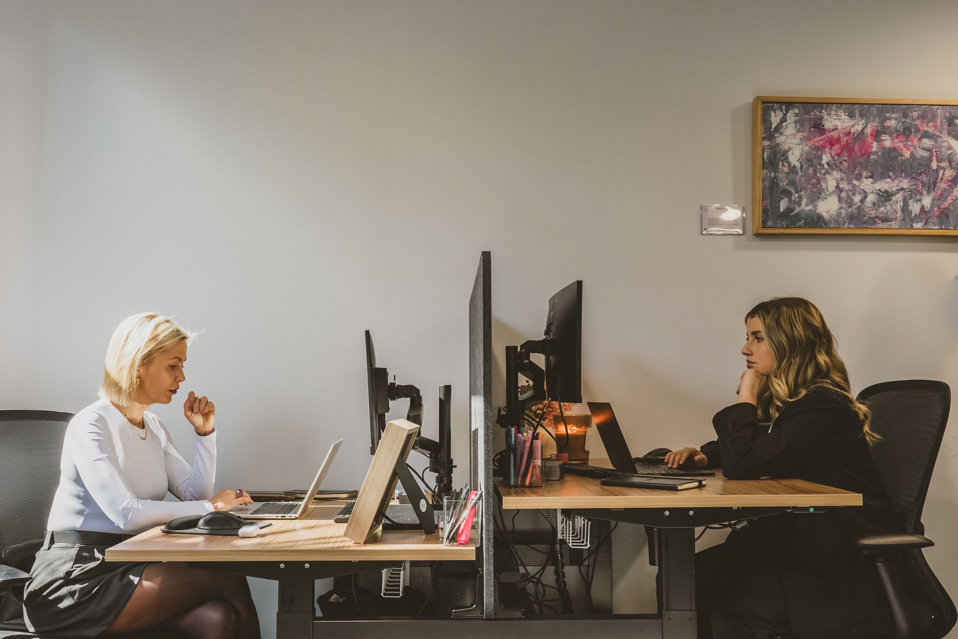 Two people working at desks in a modern office, each using a laptop, separated by a partition.