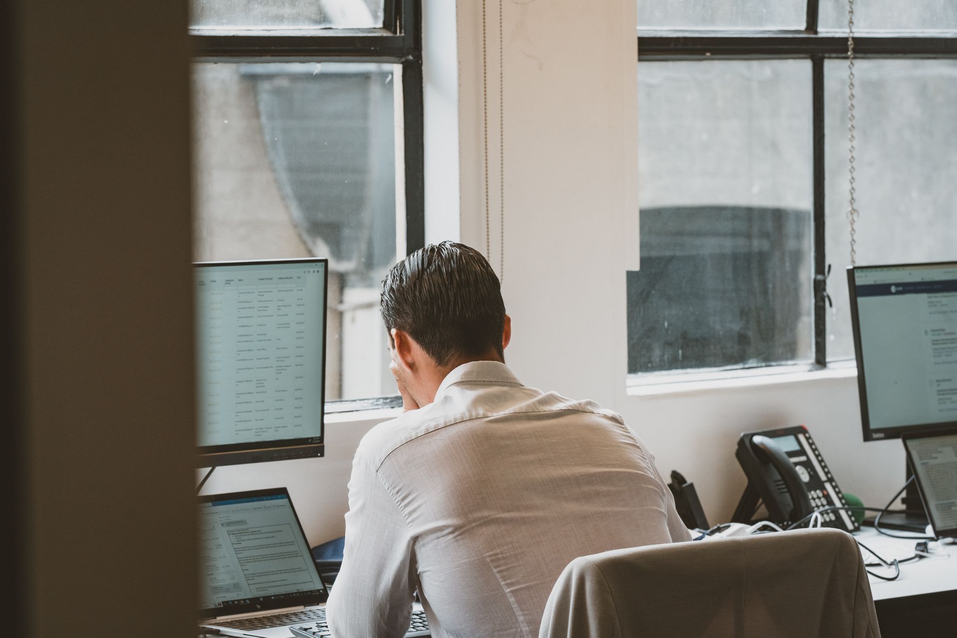 Man working at a desk with multiple computer screens, facing away from the camera in an office setting.