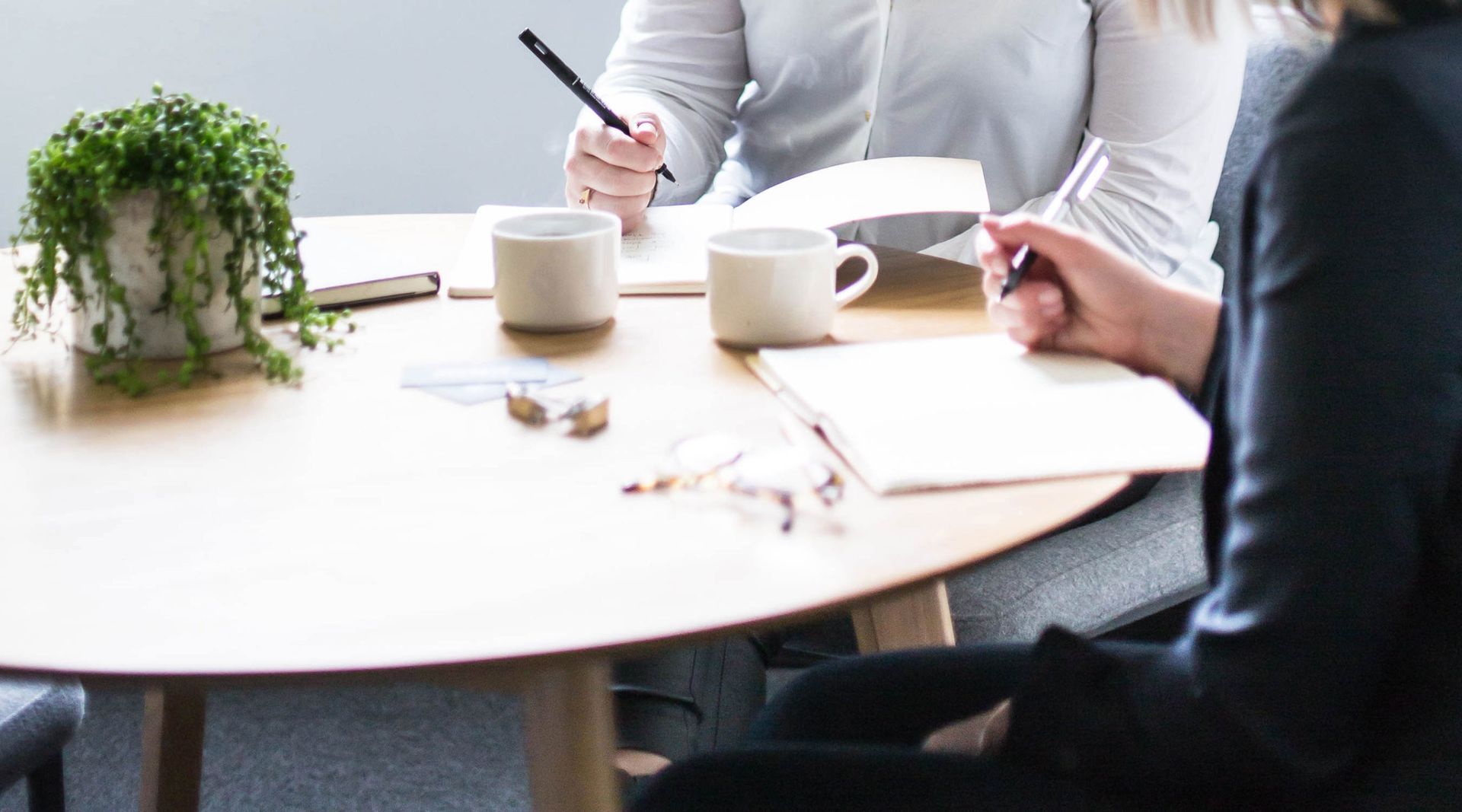 Two people at a round table, taking notes. Mugs, glasses, and a potted plant are on the table.