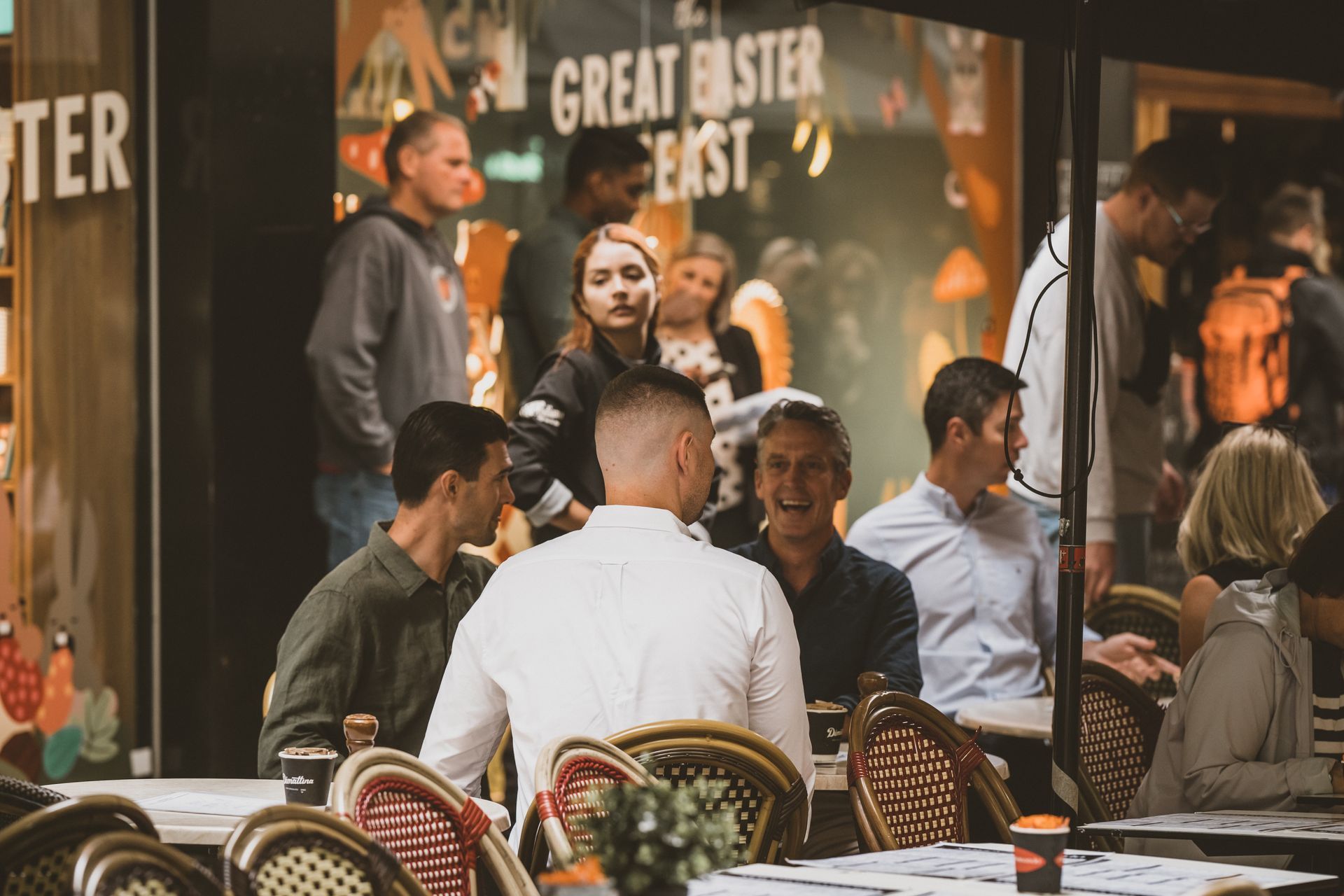 People outside a restaurant, some seated at tables, socializing and looking at each other, illuminated by outdoor lights.