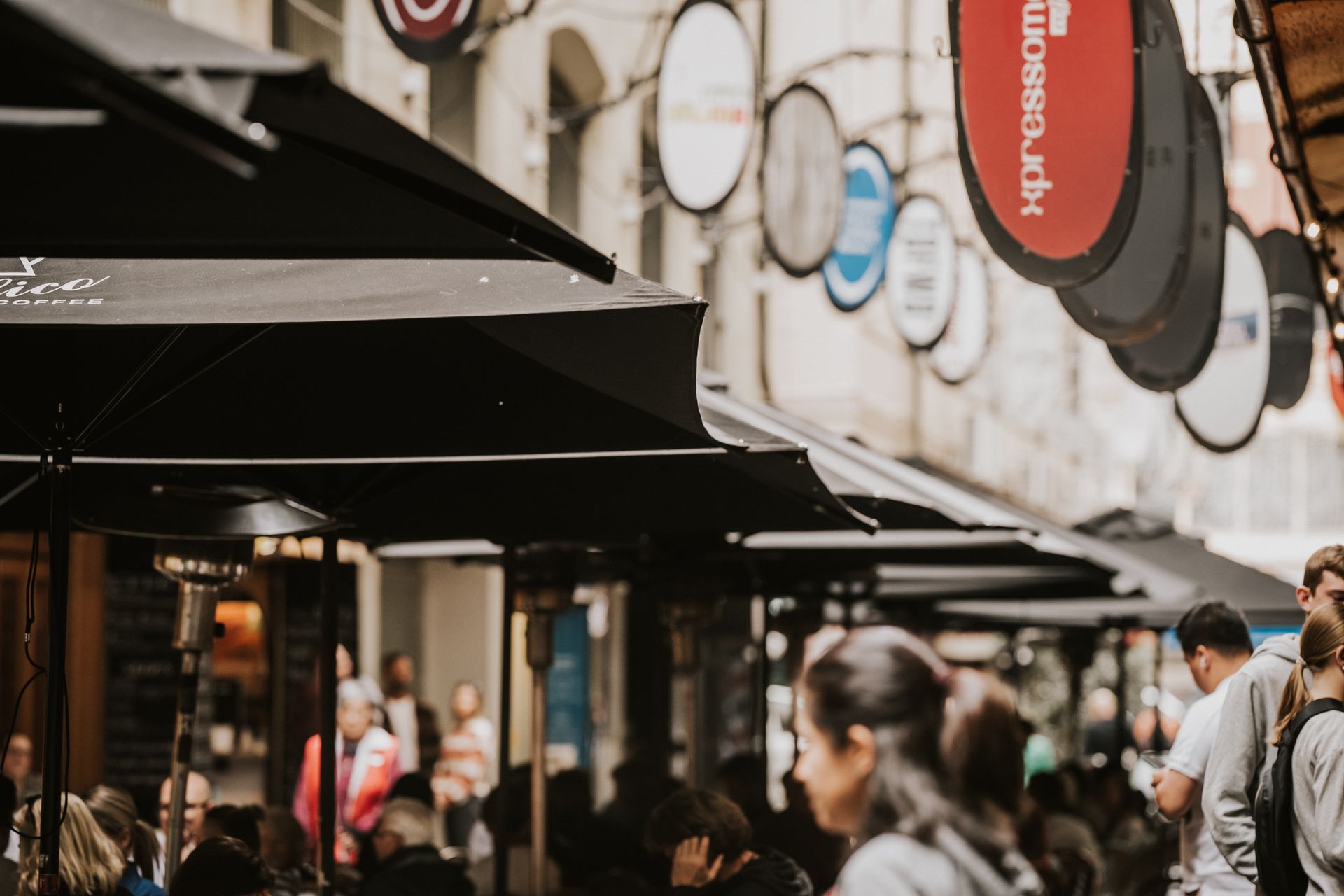 Black umbrellas shade outdoor seating area of a restaurant, street signs hang overhead. People walk by.