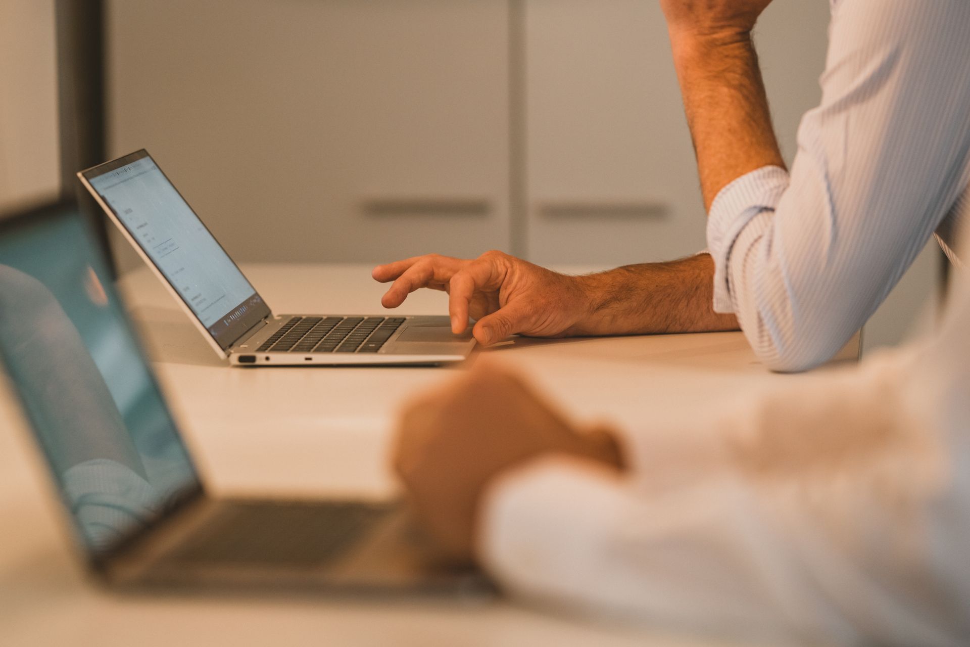 Two people working on laptops at a table. One person types on the keyboard, the other rests their chin.